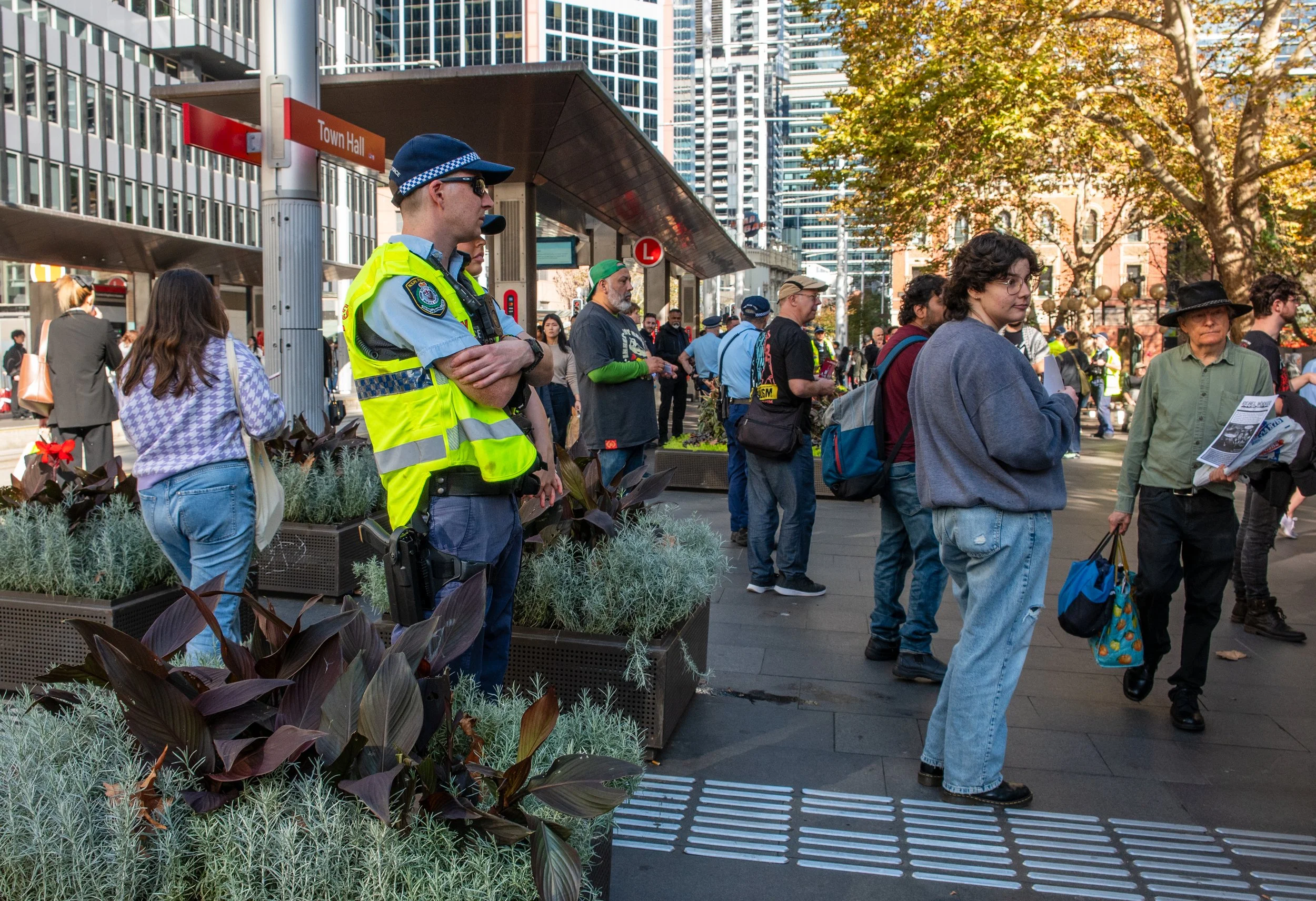 Crowd of people waiting at an outdoor city bus stop with skyscrapers in the background. A police officer in a yellow vest stands with arms crossed among the group.