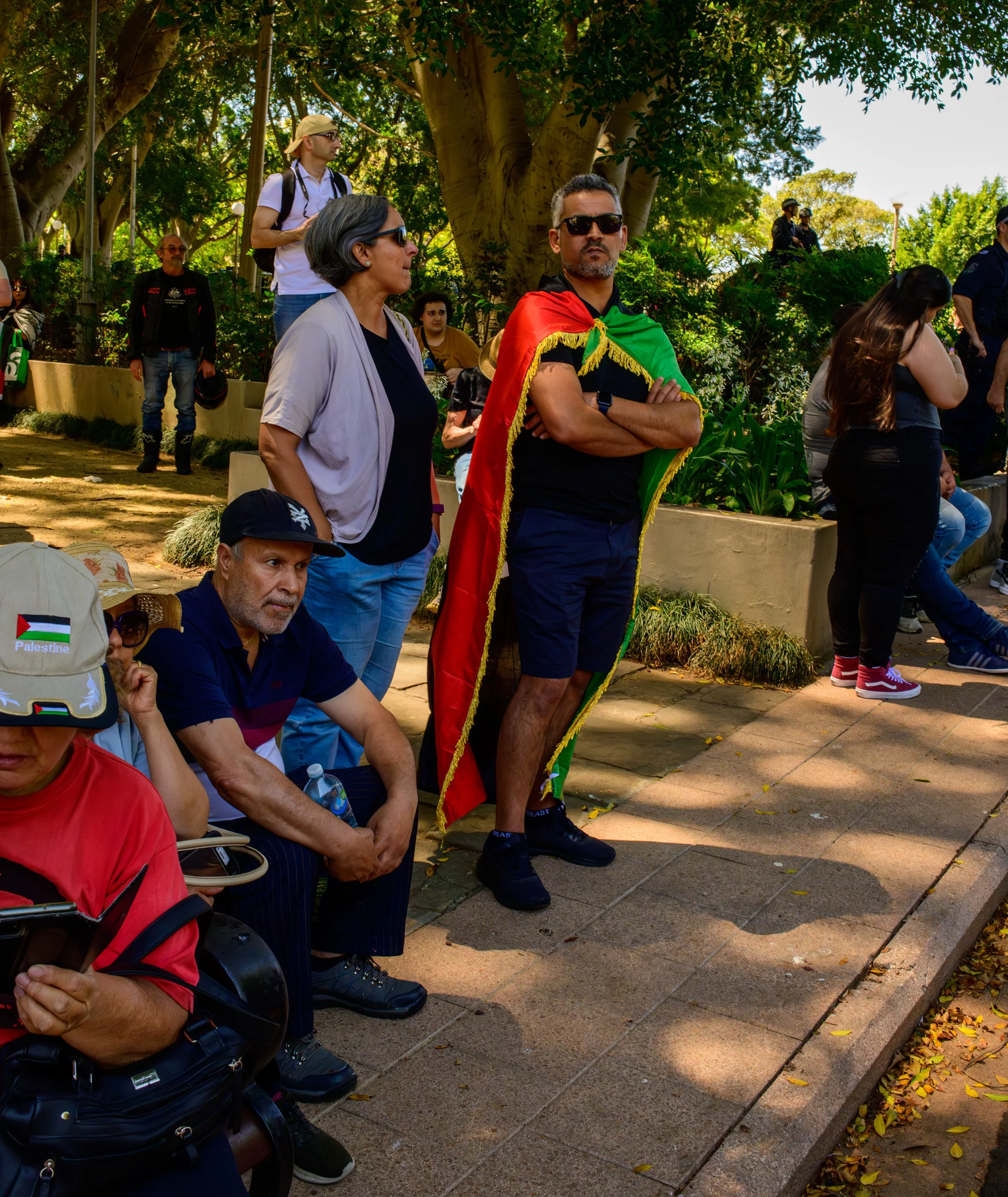 A group of people gathered outdoors in a park, with some sitting and others standing, including a man wearing a black shirt and shorts with a green, yellow, and red flag cape, and others wearing casual clothing, some with hats and sunglasses.