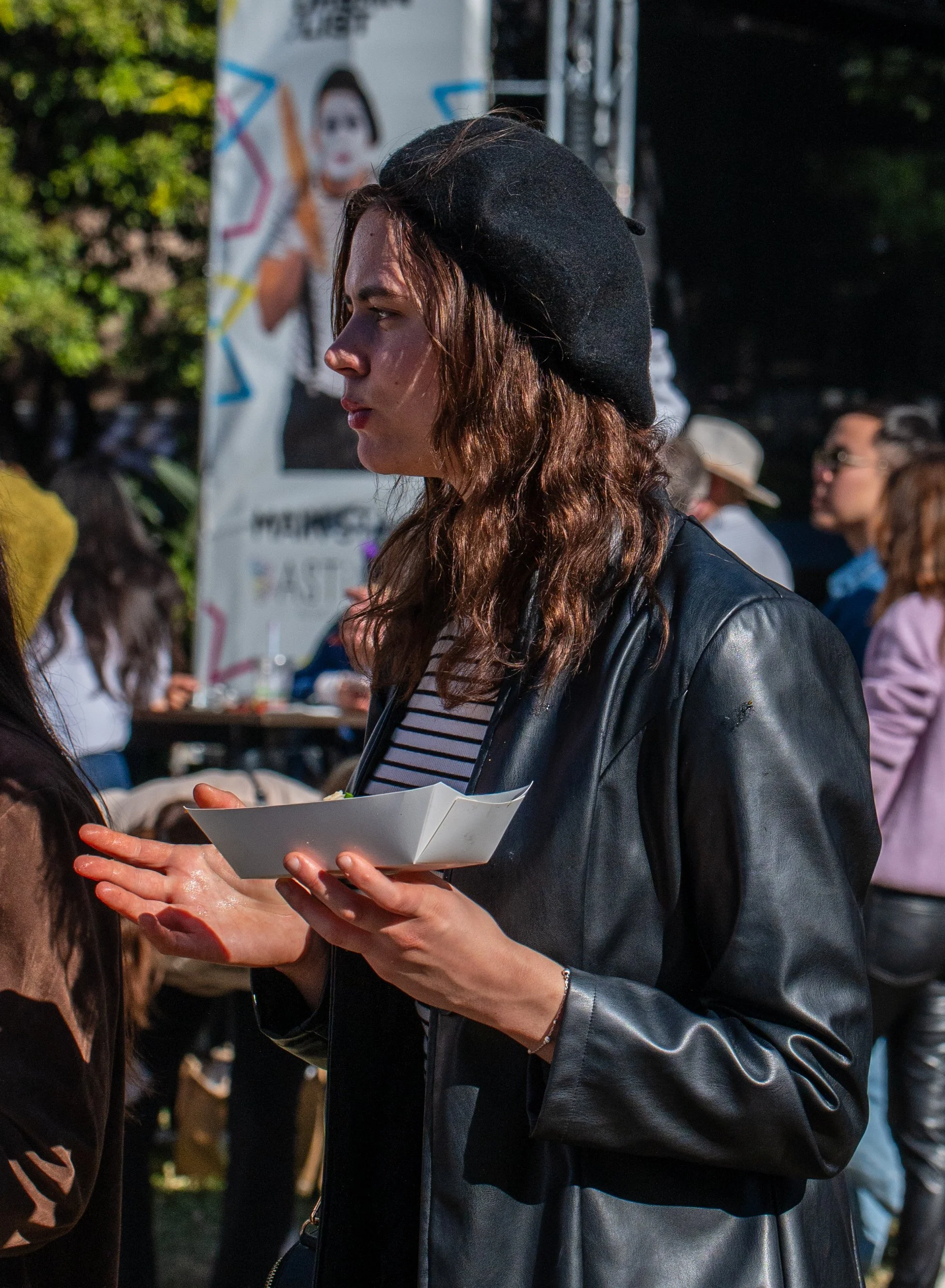 A young woman with wavy brown hair wearing a black beret, black leather jacket, and striped shirt, holding a white takeout container at an outdoor event with other people in the background.