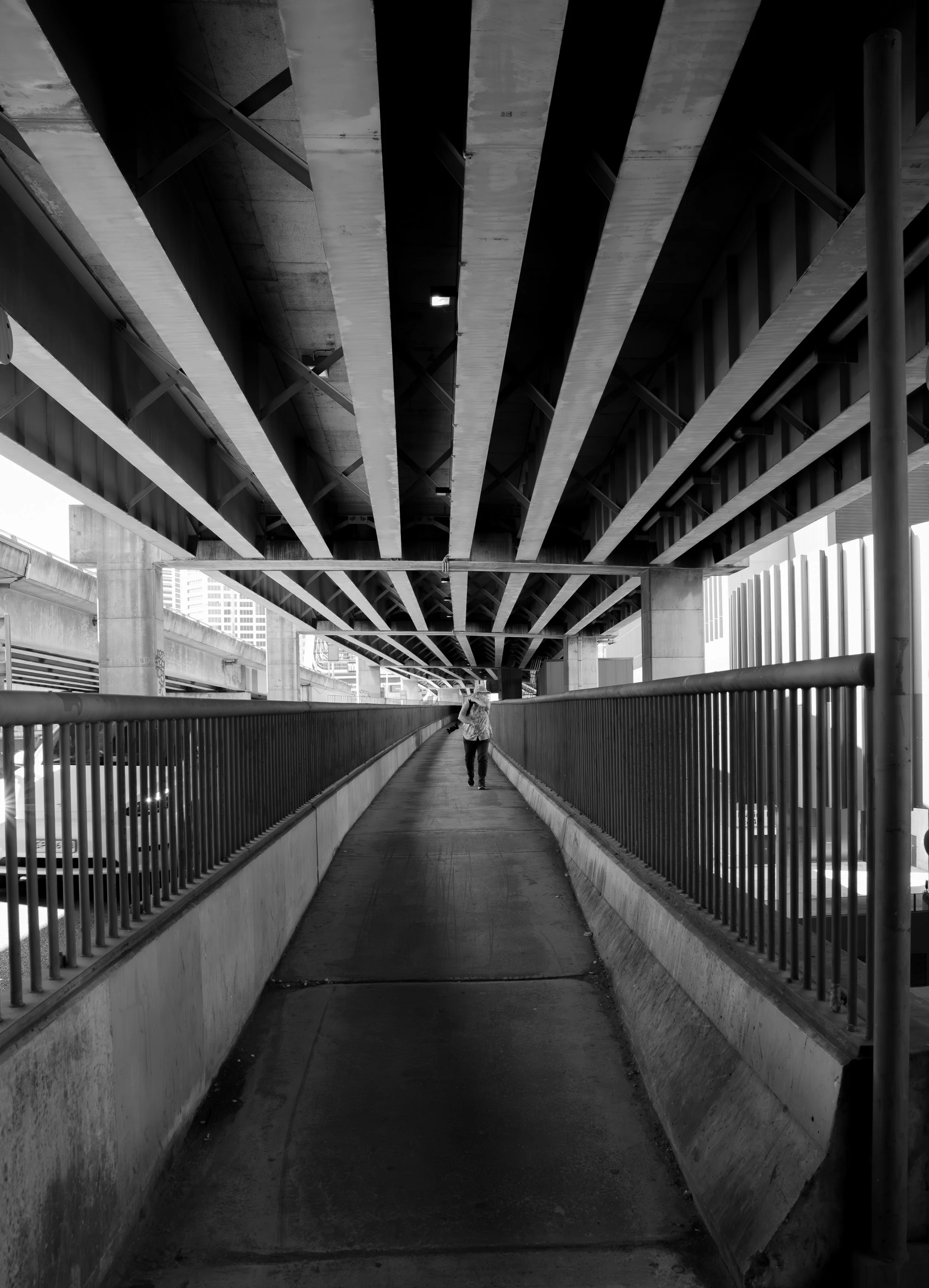 A person walking on an underpass or footbridge beneath a large multi-level highway overpass with concrete pillars and metal railings, black and white photo.