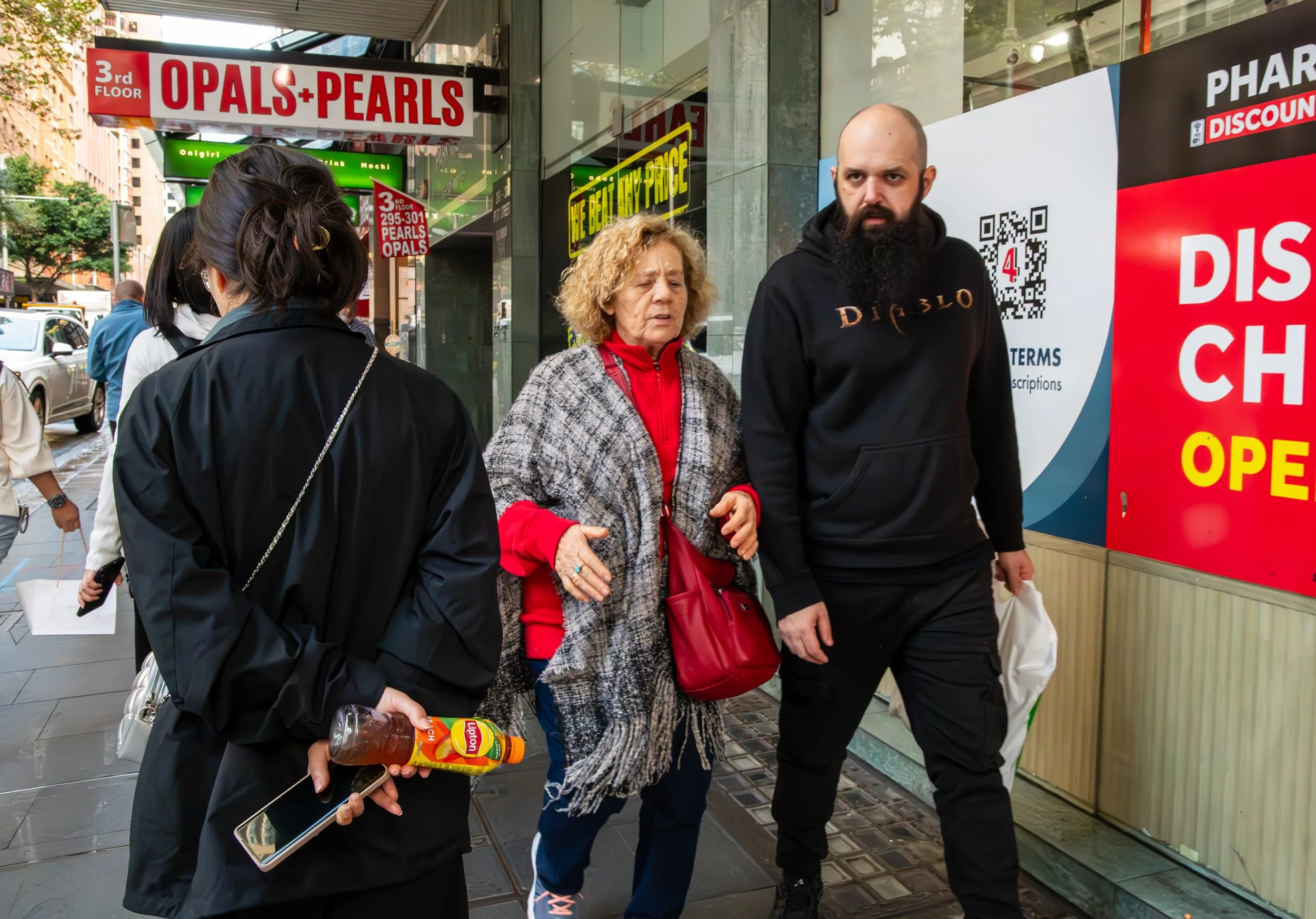 Street scene showing several people walking past store signs, including one advertising opals and pearls and another partially visible sign for a pharmacy. A woman in black holds a smartphone and a bottle of tea, while an older woman in red and gray and a man with a beard in a black hoodie walk together.