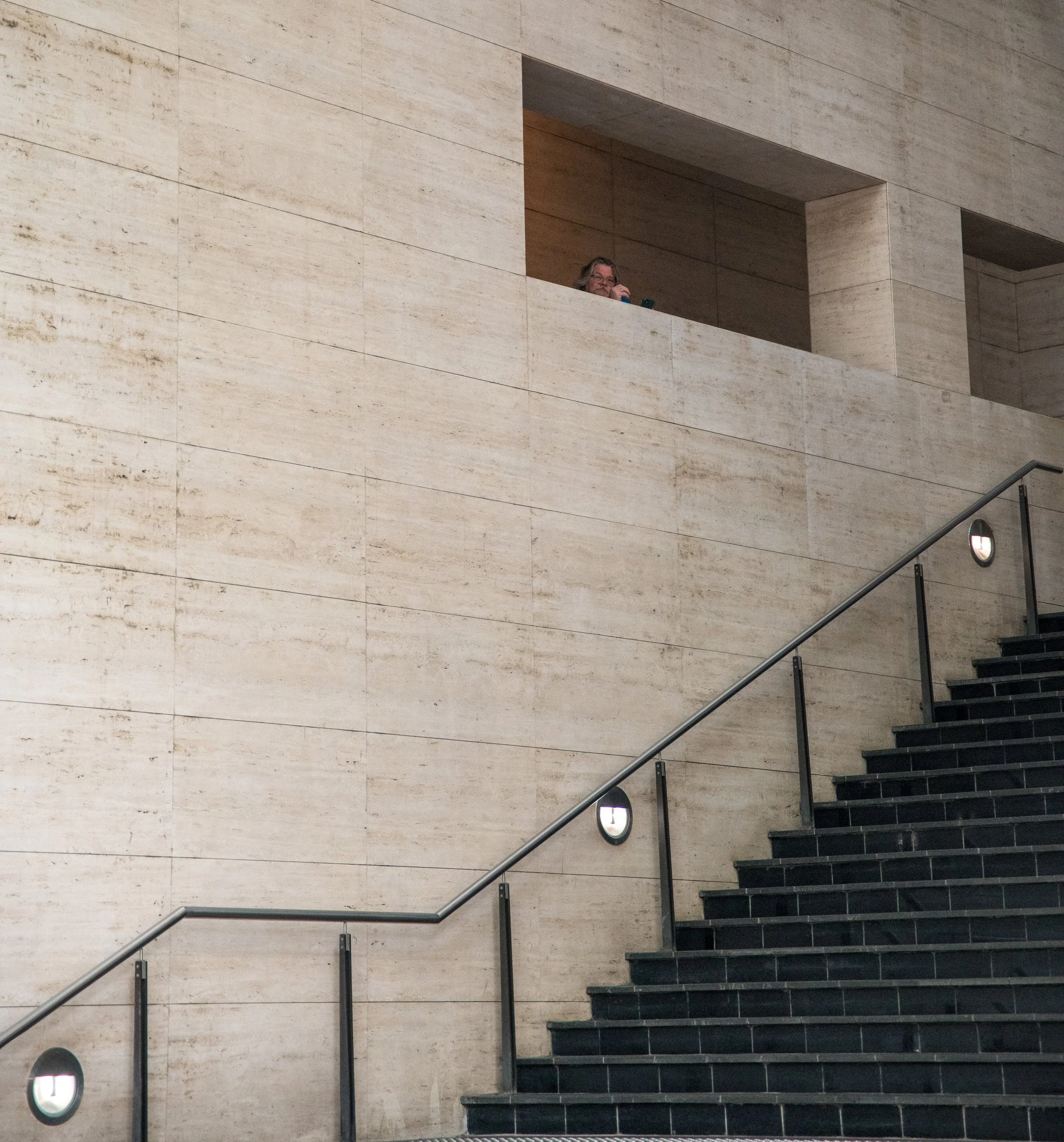 A person with glasses on a balcony looking down at the camera in a modern building with beige stone walls and black stairs.