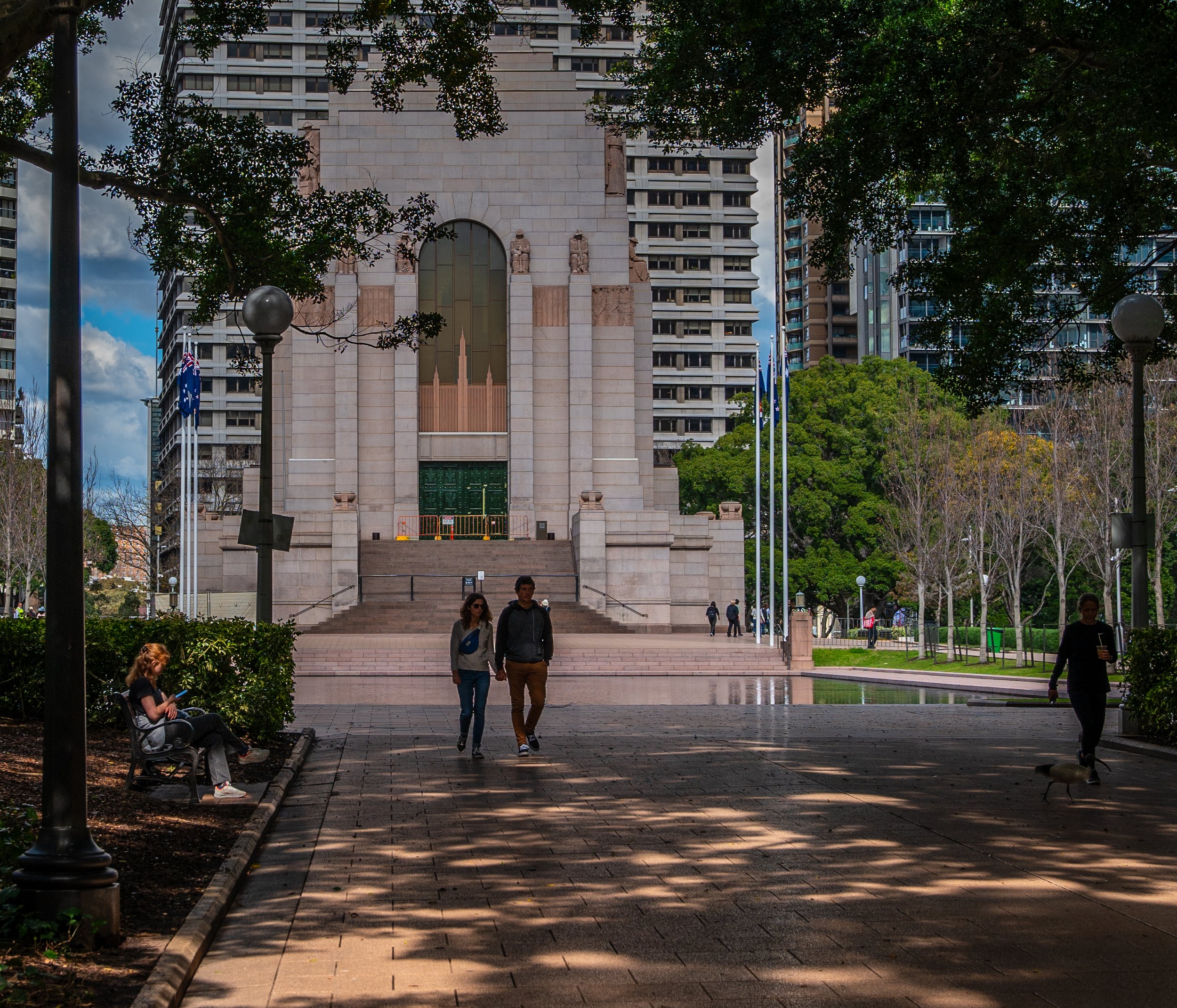 People walking and sitting on benches in a city park with tall buildings and trees in the background.