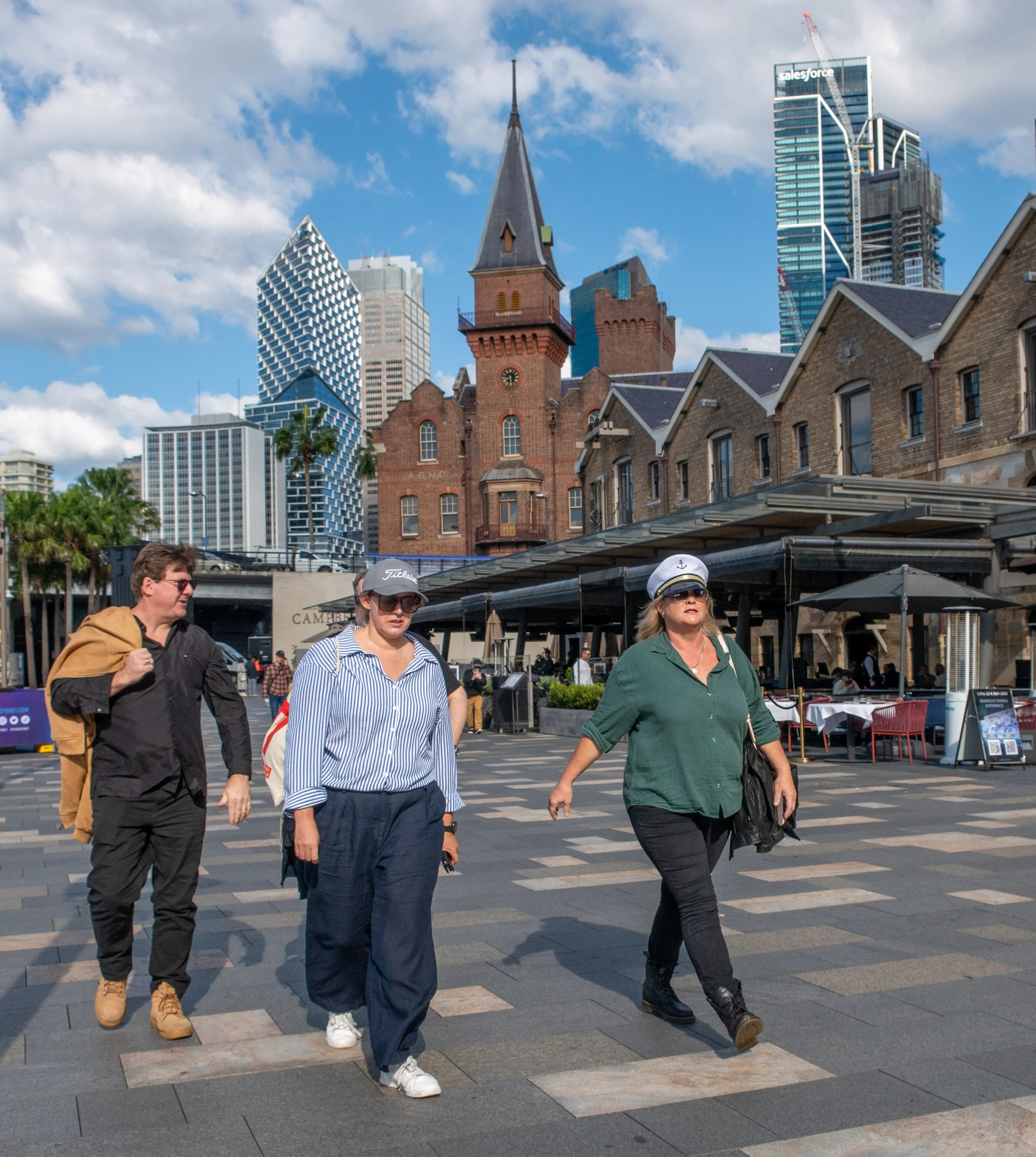 Three people walking across a city street with modern skyscrapers and historic brick buildings in the background on a sunny day.