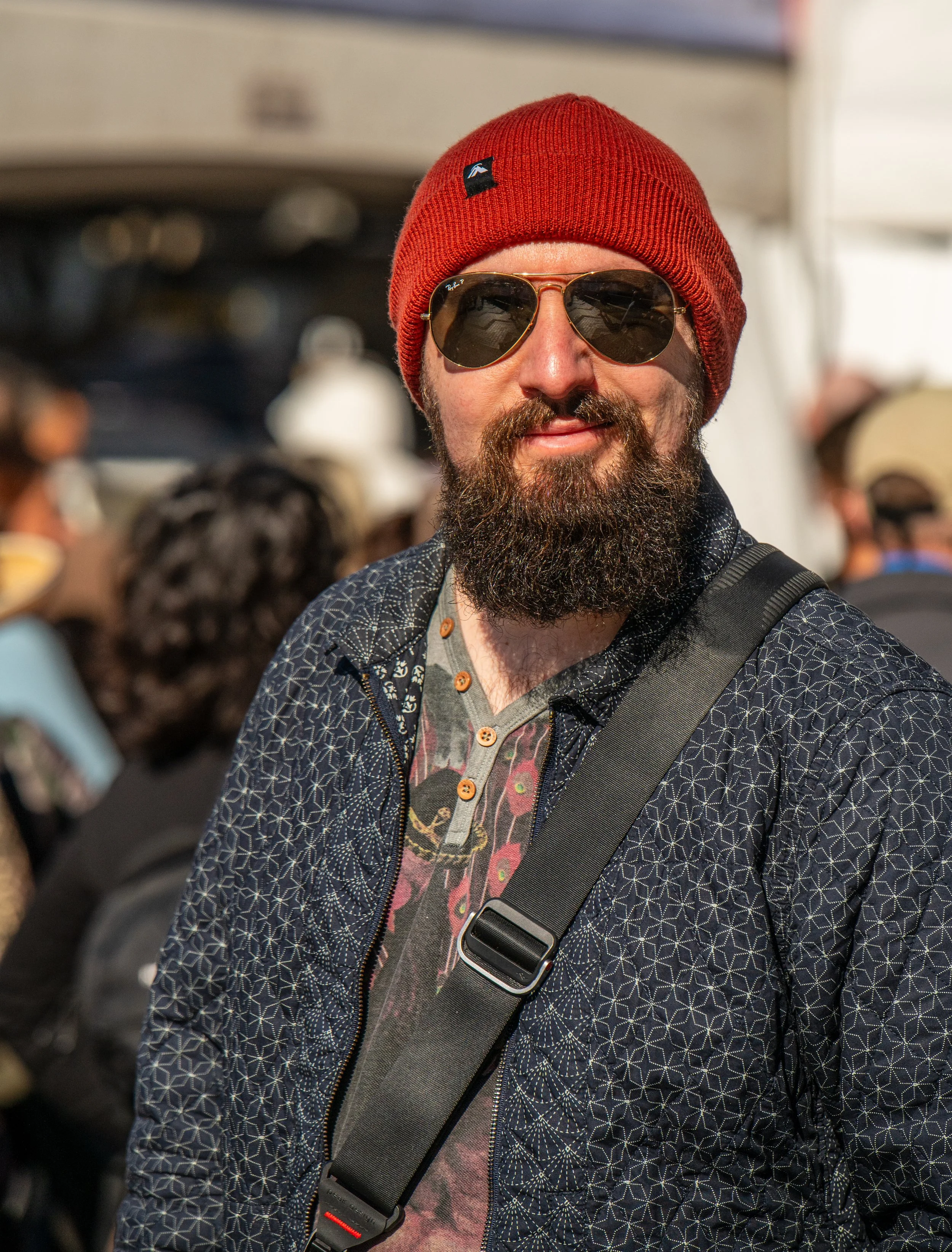 Man with a beard wearing sunglasses, a red beanie, a patterned jacket, and a crossbody bag at an outdoor event.