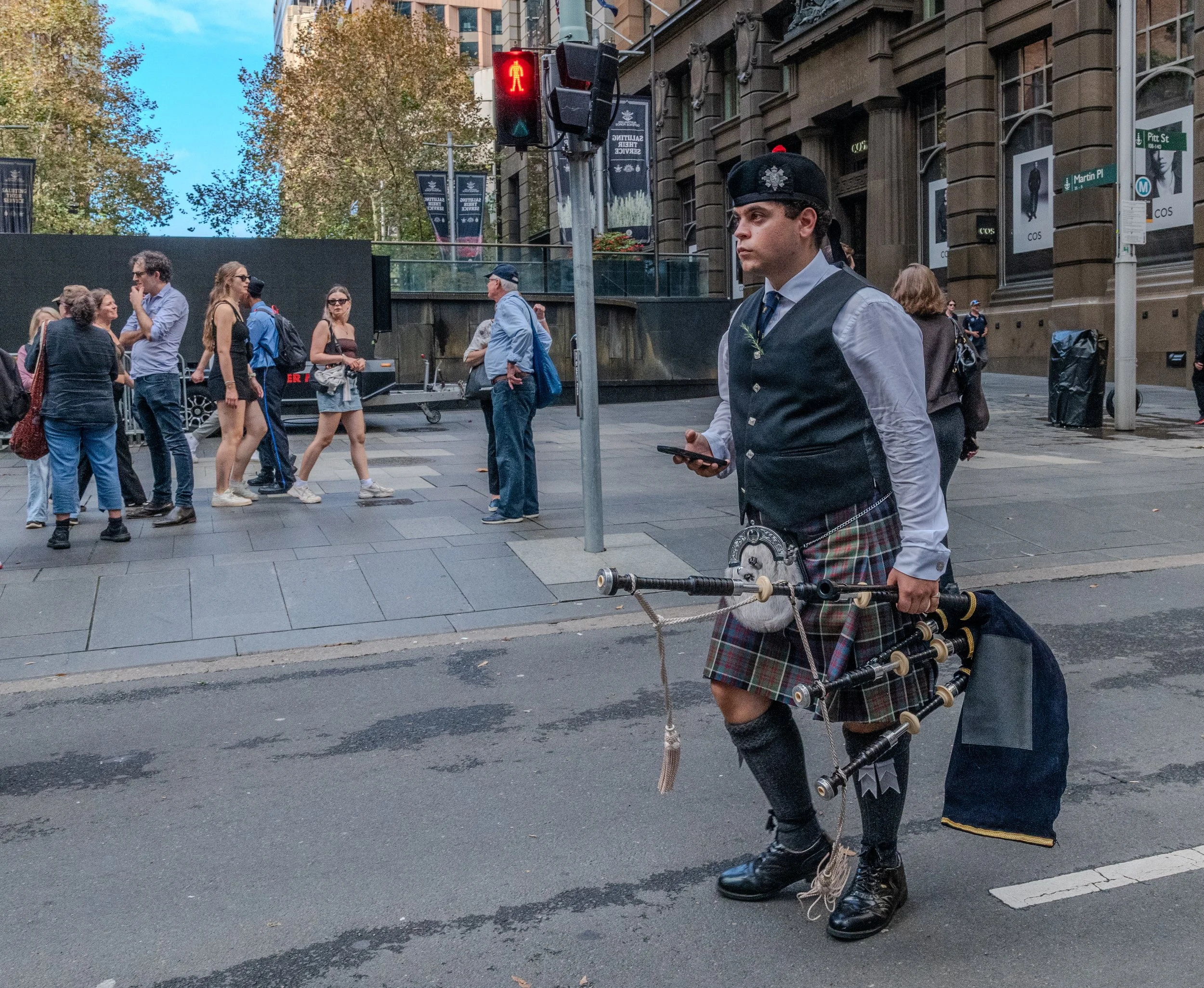 A man dressed in traditional Scottish attire, including a kilt and a sporran, stands on a city street holding a set of bagpipes while looking at his phone. People are walking and waiting to cross at a pedestrian signal. A red pedestrian crossing light is visible above him.
