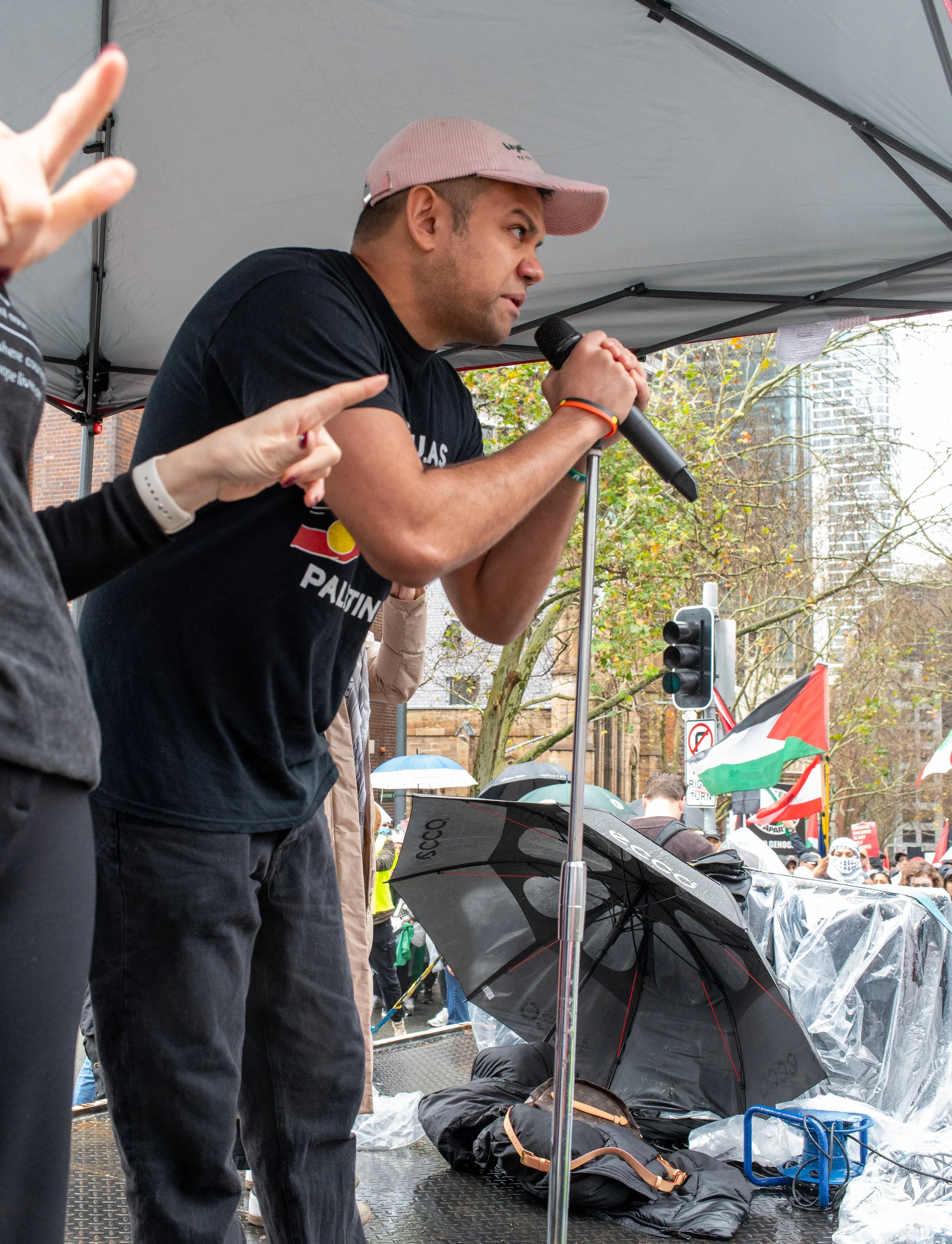 A man speaking into a microphone at an outdoor protest or rally, with a crowd holding flags and umbrellas in the background.