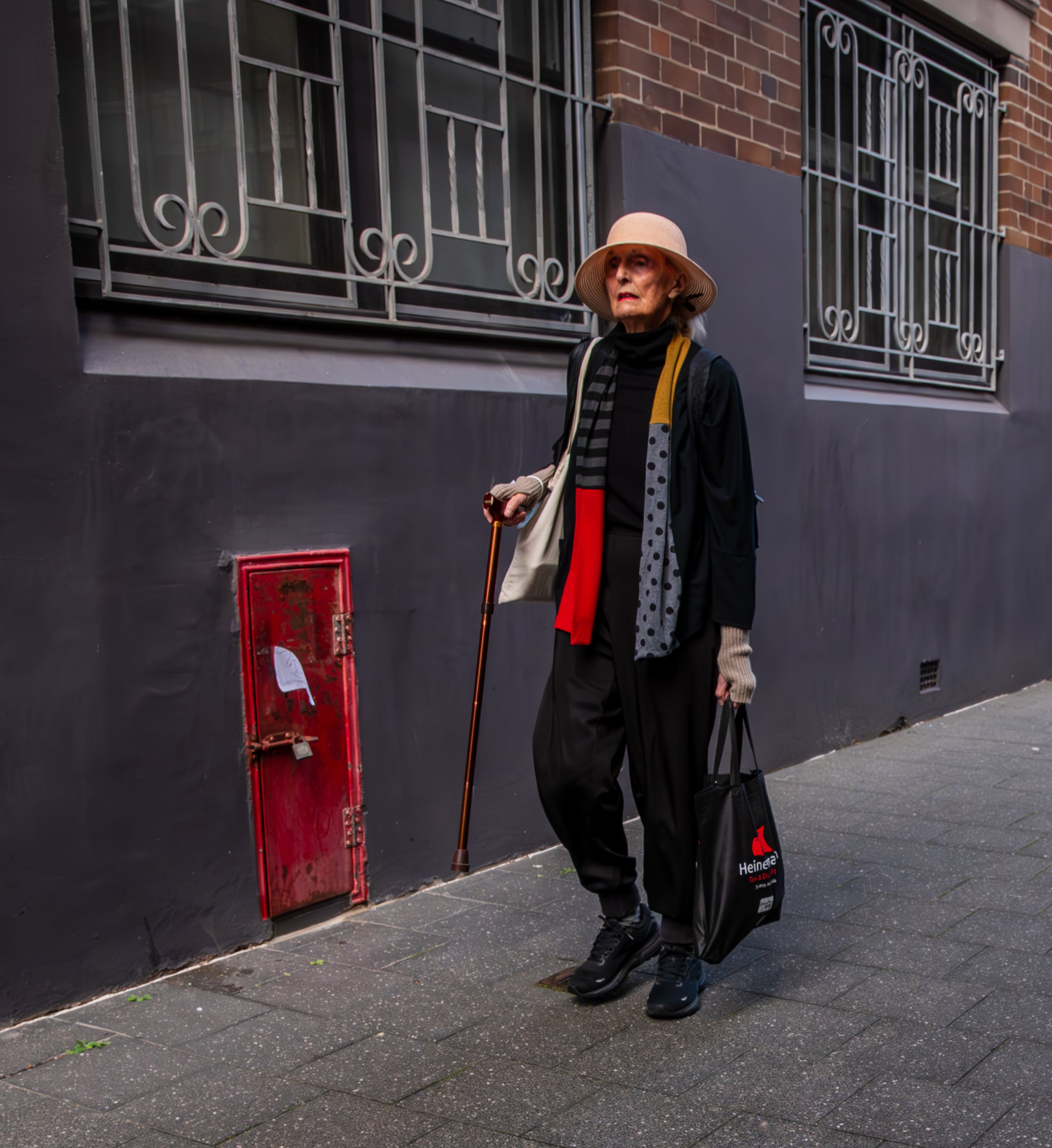 An elderly woman wearing a wide-brimmed hat, black pants, and a black jacket, standing on a sidewalk against a dark wall with windows and decorative iron railings, holding a cane, a shopping bag, and a tote bag
