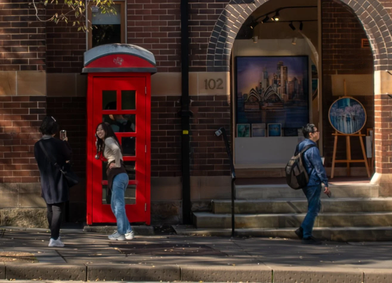 Three people outside an art gallery with a red telephone booth in front, one taking a photo of the other, and an art display inside the gallery visible through the entrance.