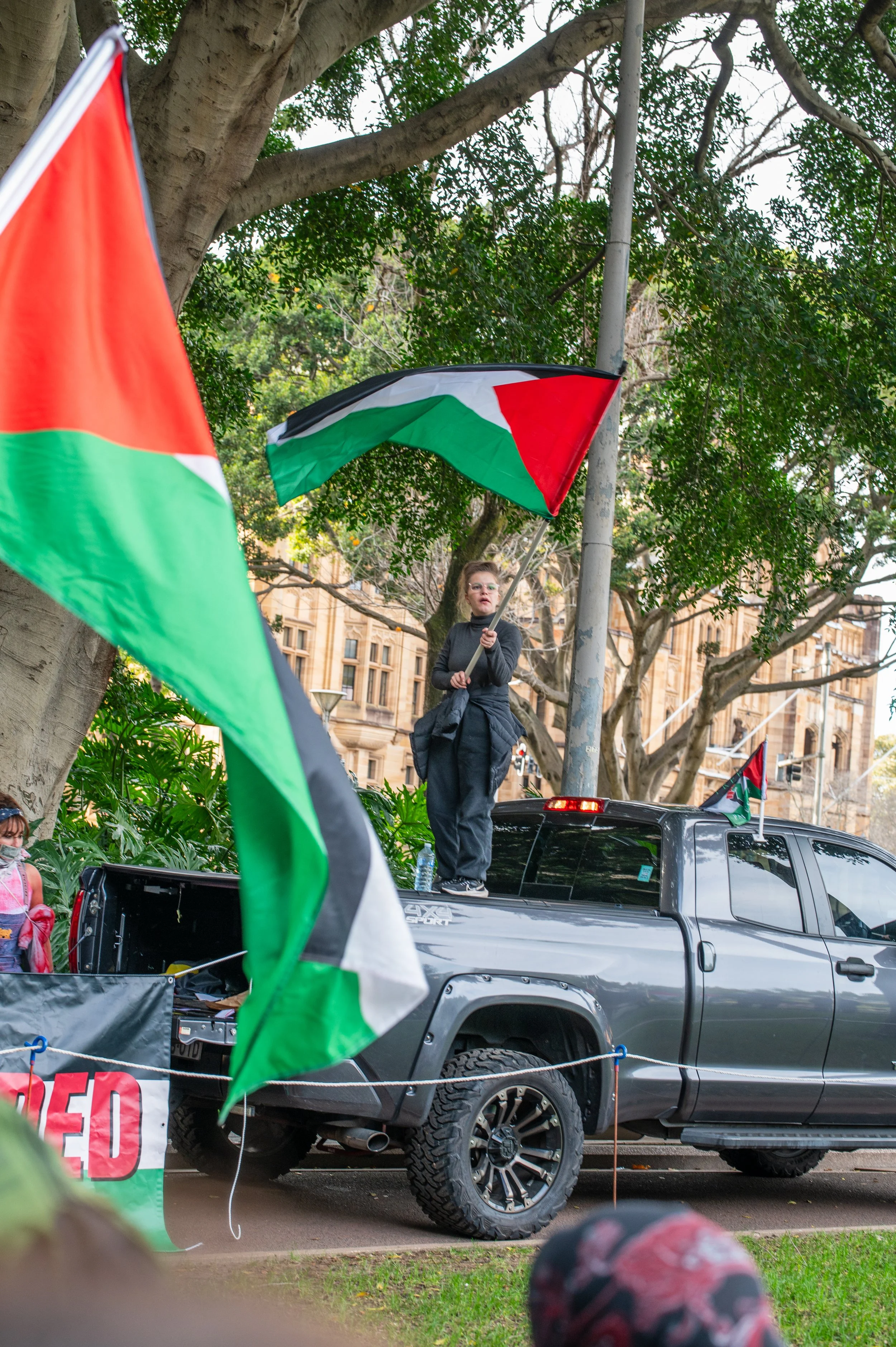 A woman standing on the back of a gray pickup truck holding a Palestinian flag during a protest or demonstration.