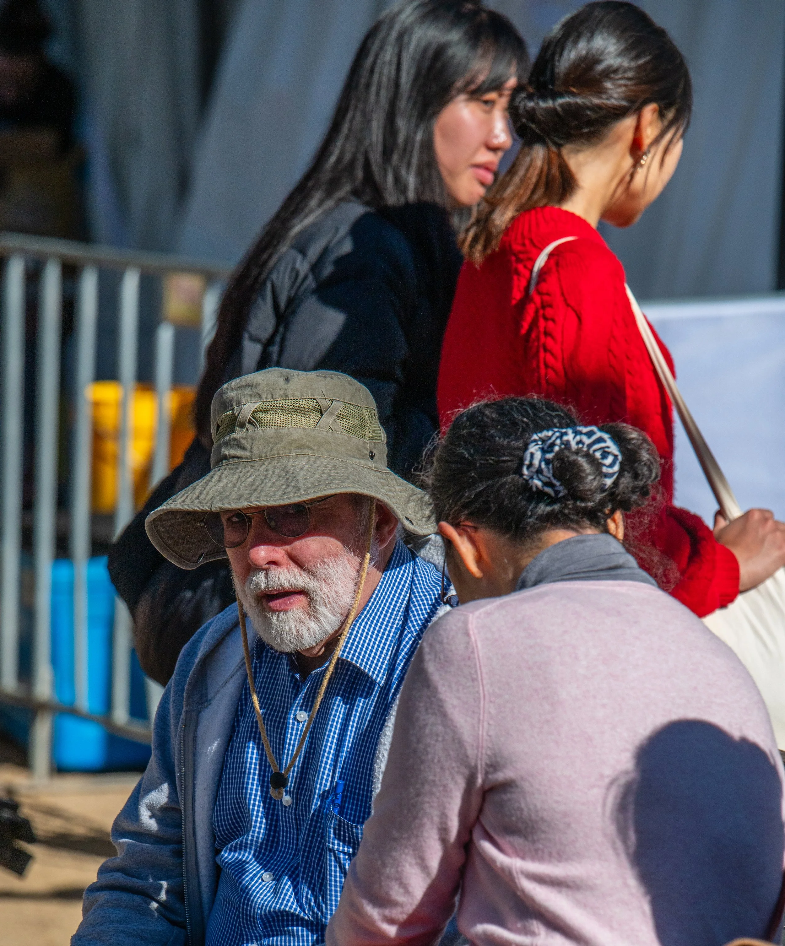 A group of people sitting outdoors, including an elderly man with a wide-brimmed hat, glasses, and a blue checkered shirt, engaged in conversation.