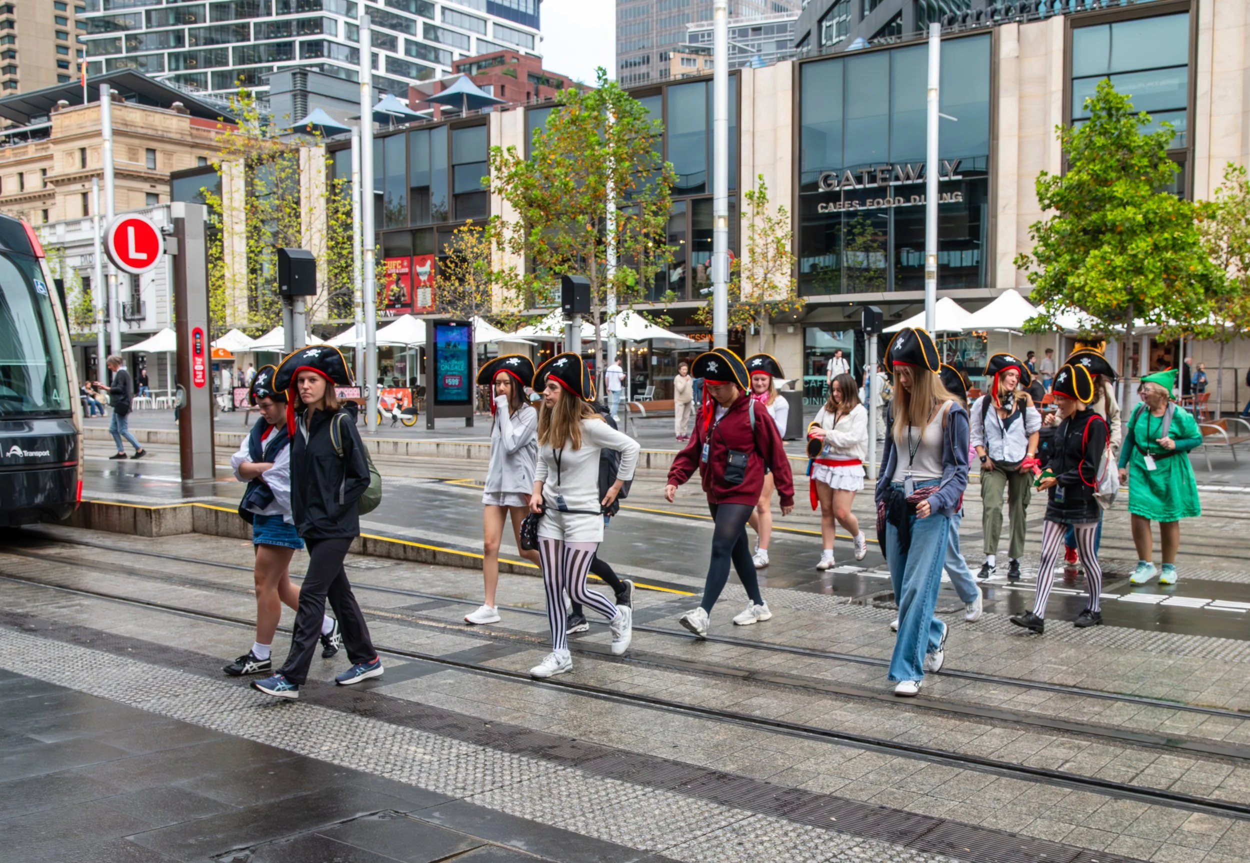 Group of people dressed as pirates walking across a city street with a tram track, modern buildings, and a shopping center in the background.