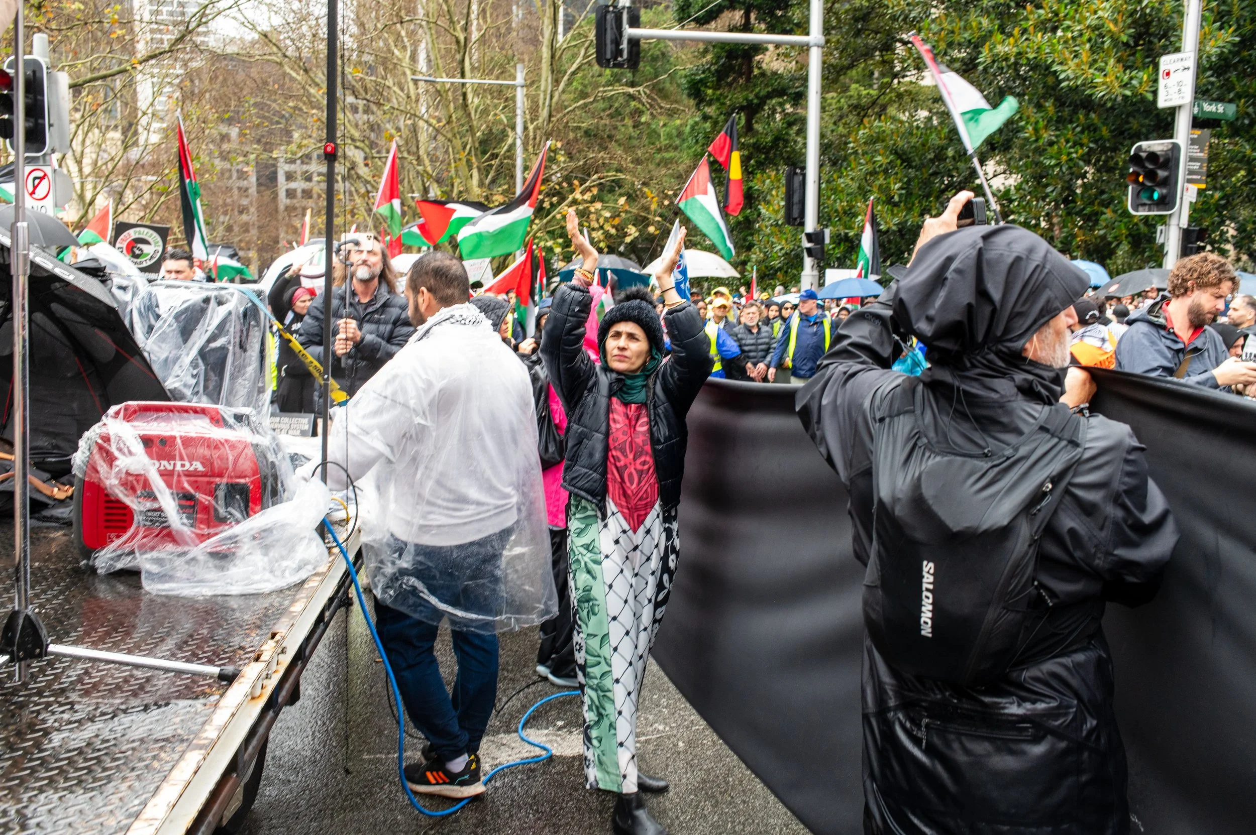 Protesters holding Palestinian flags during a demonstration on a rainy day in an urban area, with some people taking pictures and others holding signs.