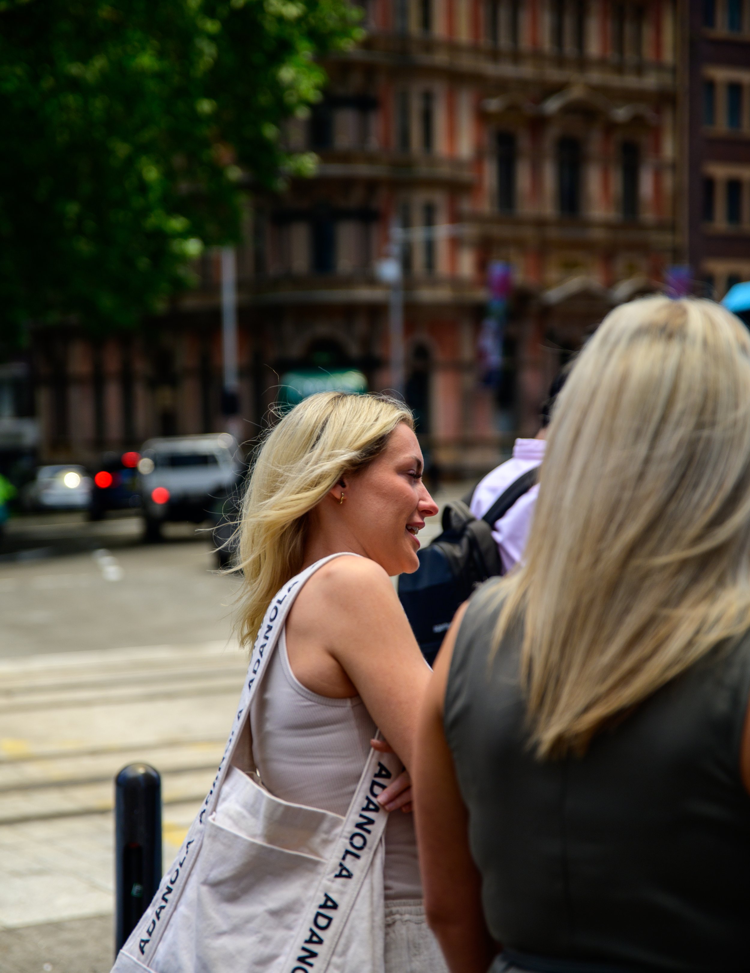 A woman with blonde hair smiling and talking outdoors on a city street, with a building in the background and other people nearby.