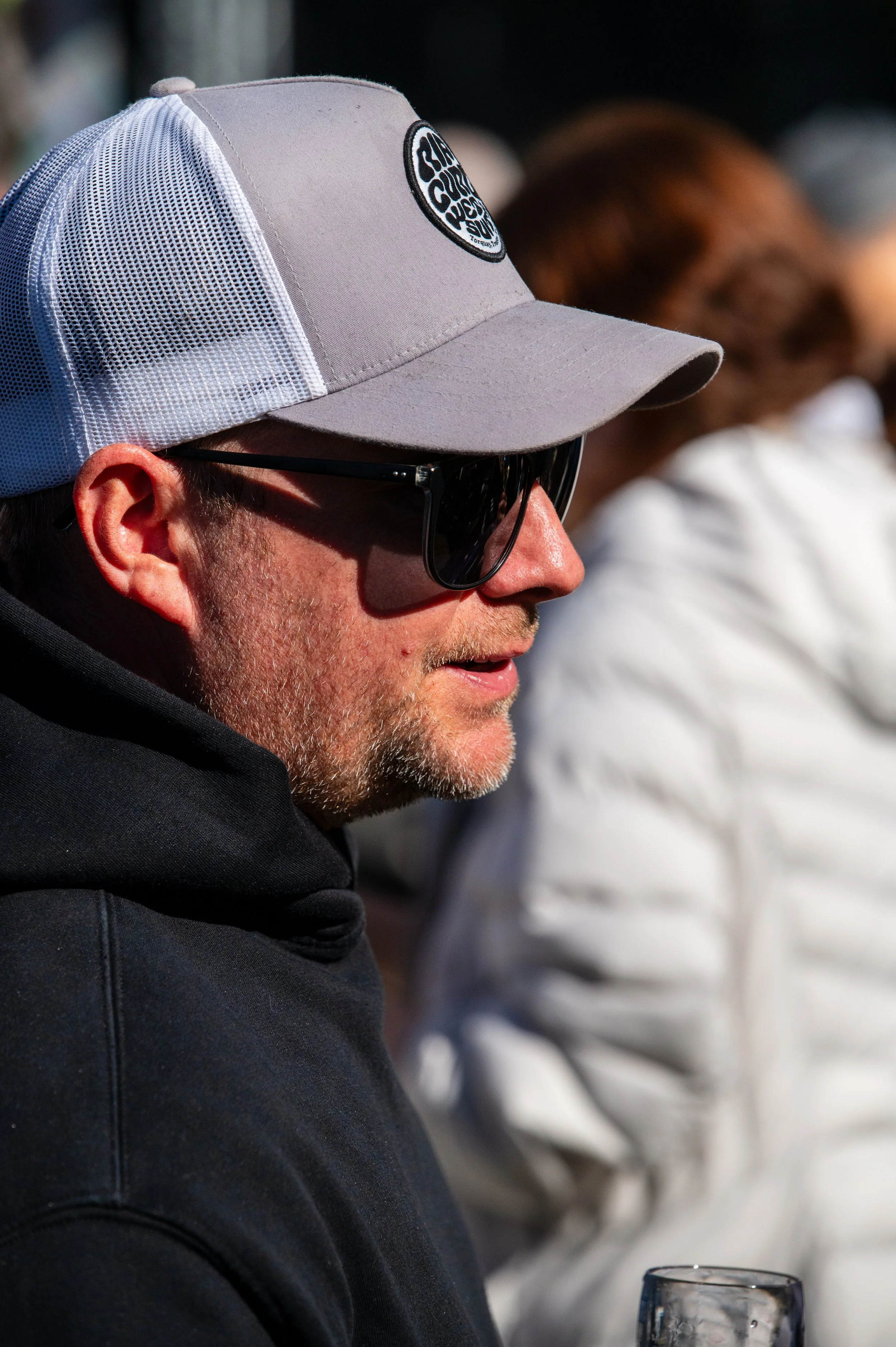 A man wearing a gray cap with a mesh back, black sunglasses, and a black hoodie, sitting outdoors with a woman in a white jacket blurred in the background.