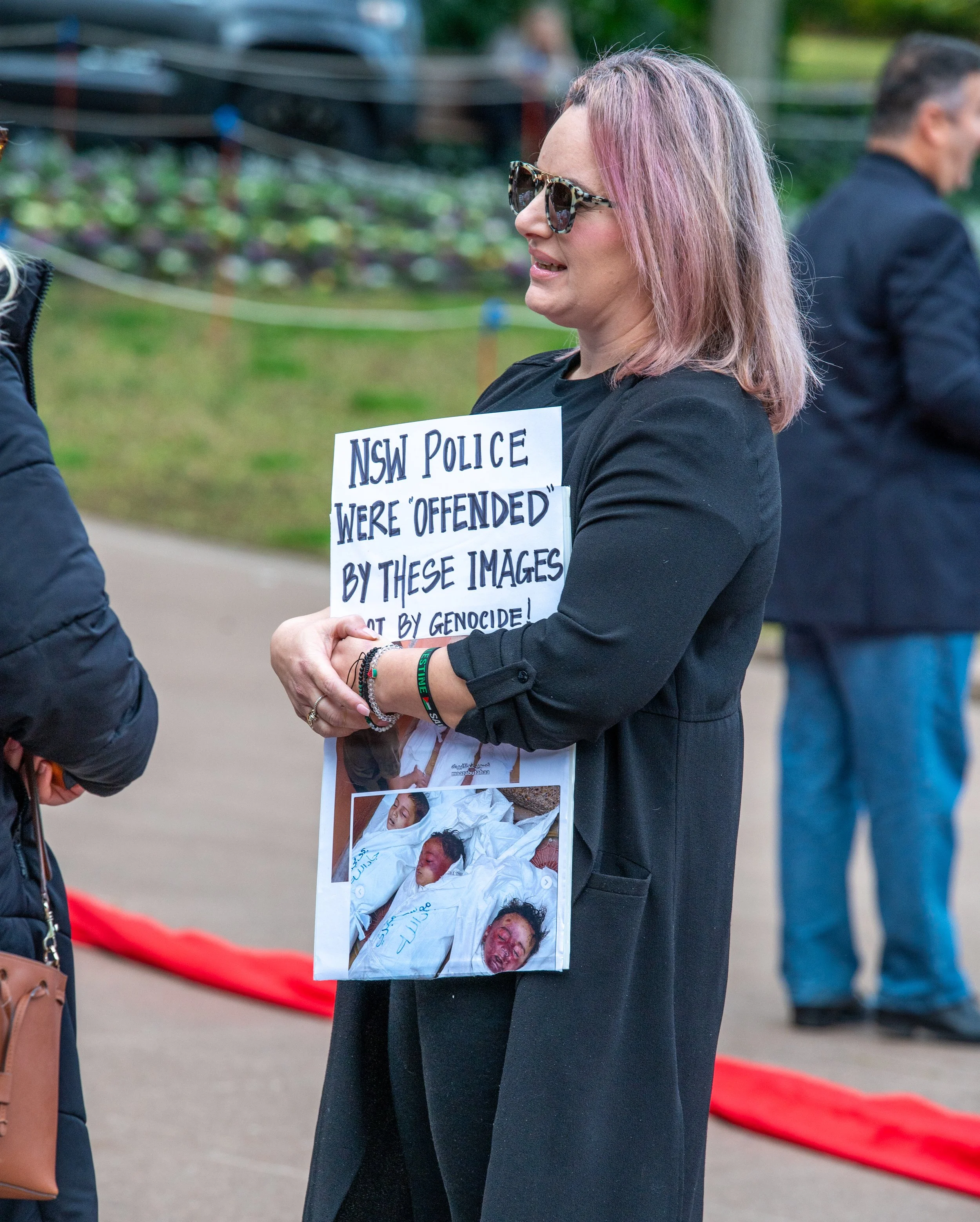 A woman protesting at an outdoor event, holding a sign with images of injured or deceased people, and text criticizing NSW police for being offended by the images.