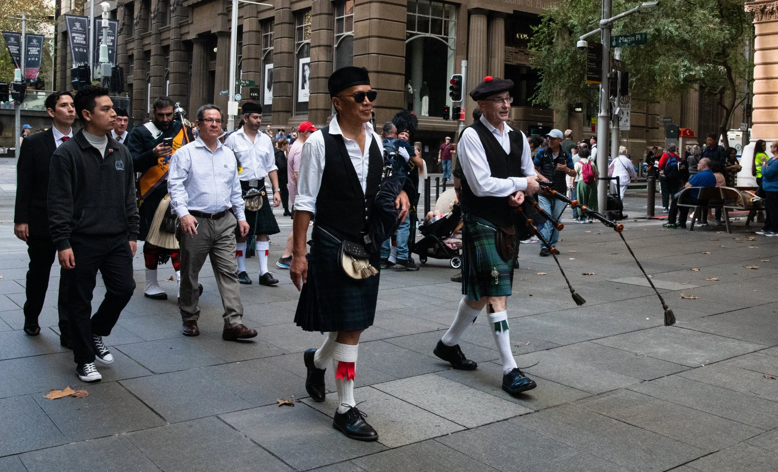 People walking and playing bagpipes on a city street with buildings and trees in the background.