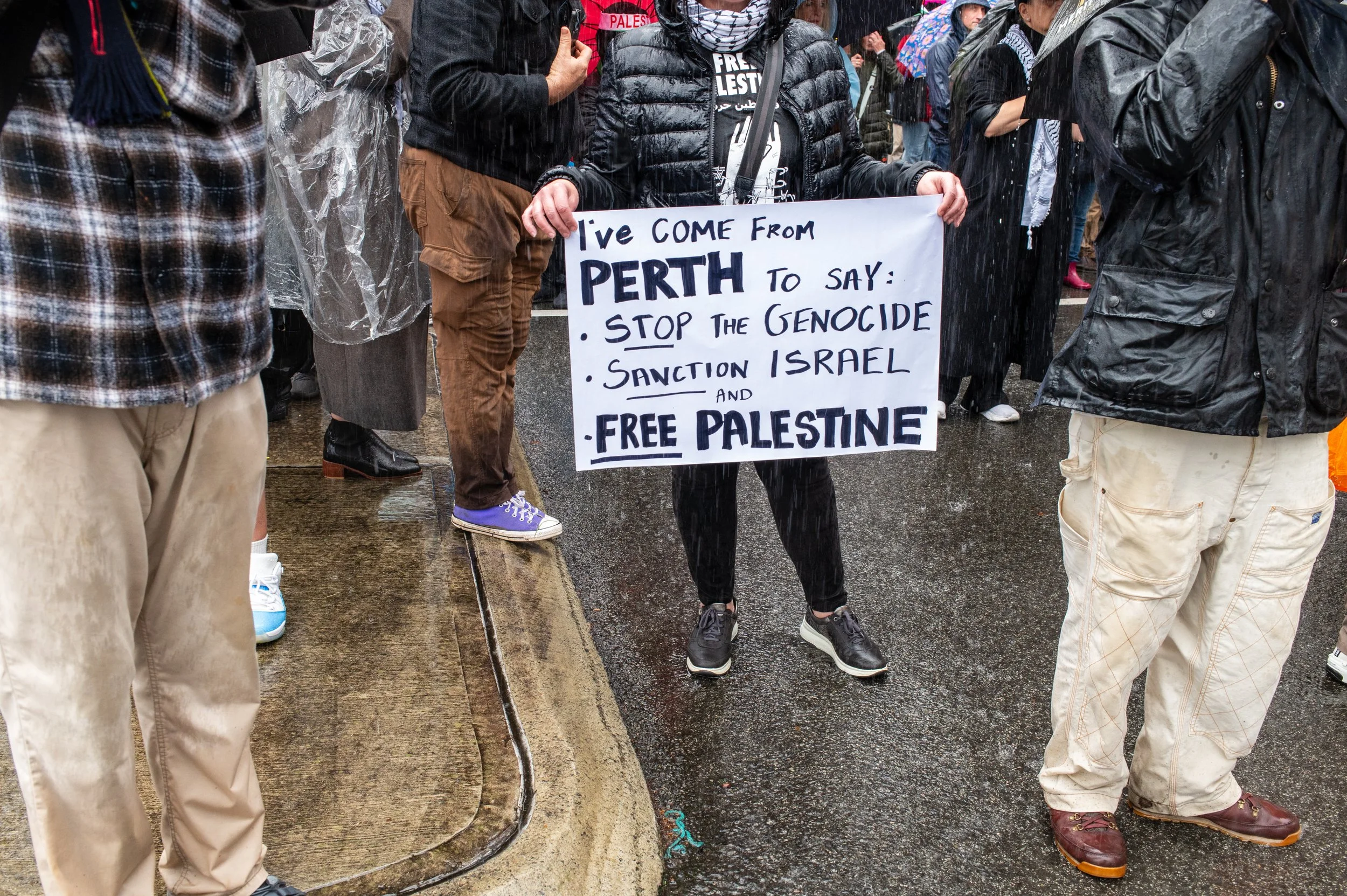A person holding a sign at a protest that says 'I've come from Perth to say: Stop the genocide, Sanction Israel, and Free Palestine.' The scene is rainy, and there are other protesters around, some wearing rain gear and holding umbrellas.