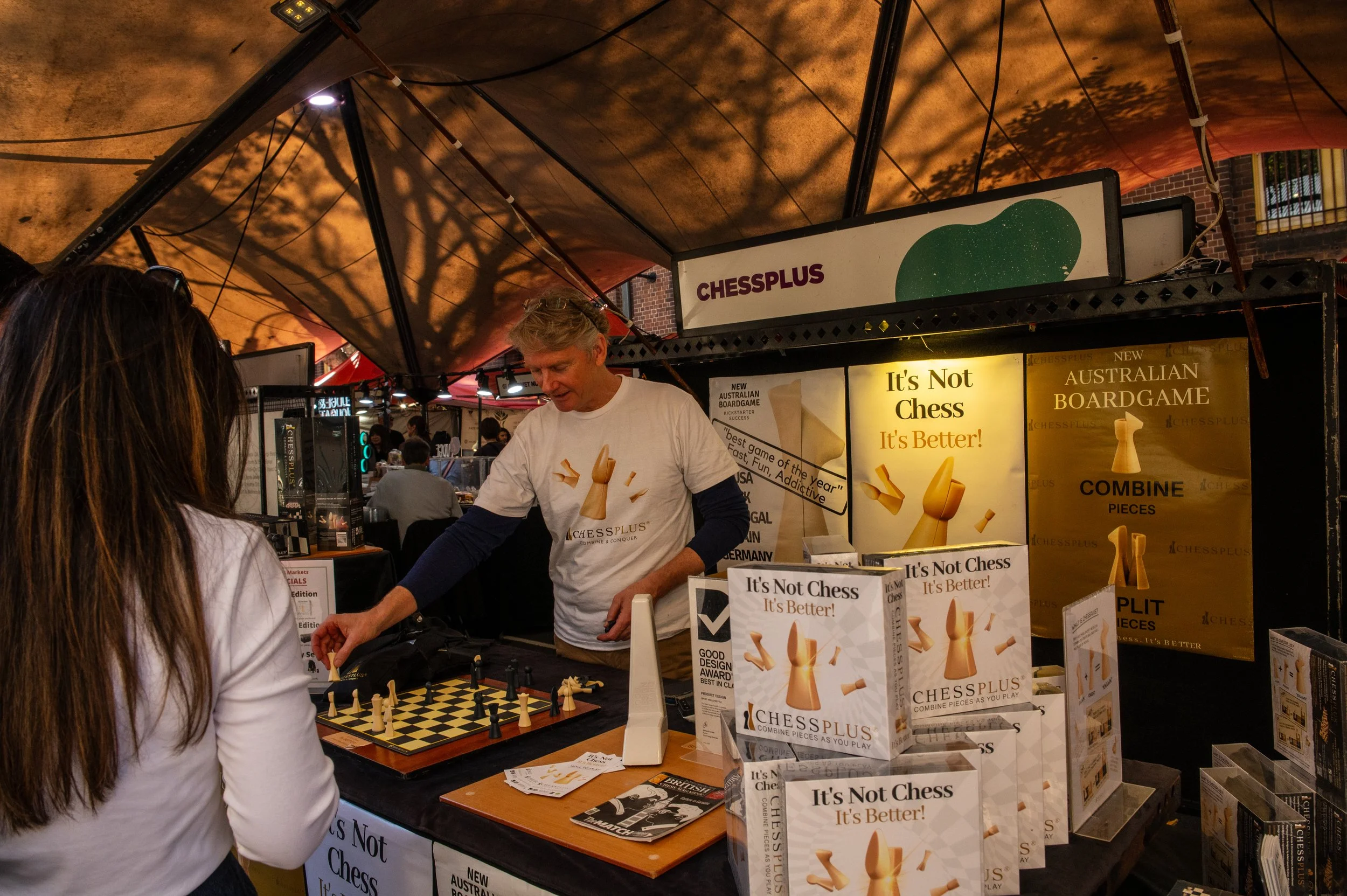 A man demonstrating a chessboard game called ChessPlus at a booth in an indoor event space. A woman is watching the demonstration, and there are promotional posters and boxes of the chess game displayed on the table. The booth has a sign that reads 'CHESSPLUS' and a large poster stating 'It's Not Chess, It's Better!'