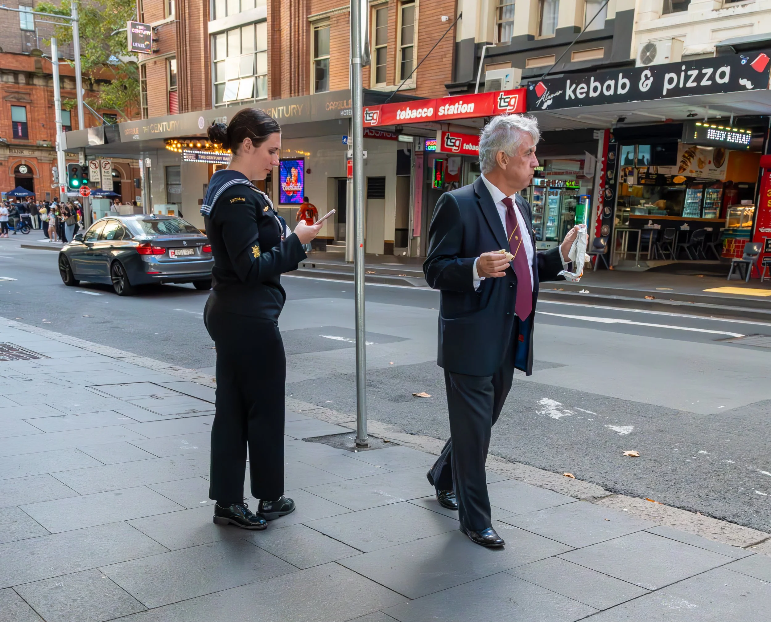 A woman in a black uniform is standing on a street corner looking at her phone, while a man in a suit and tie is eating and holding a paper bag, on a city street with shops and cars in the background.