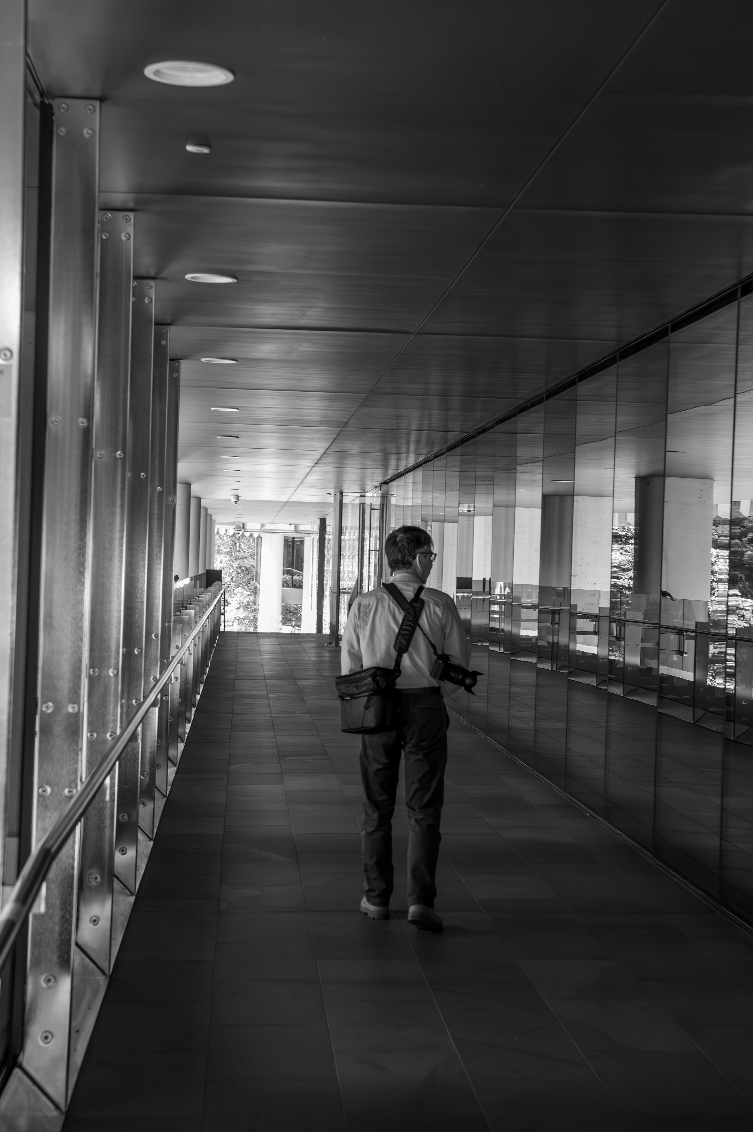A man walking down an empty corridor with reflective glass walls and modern architectural design, carrying a camera and wearing a backpack.