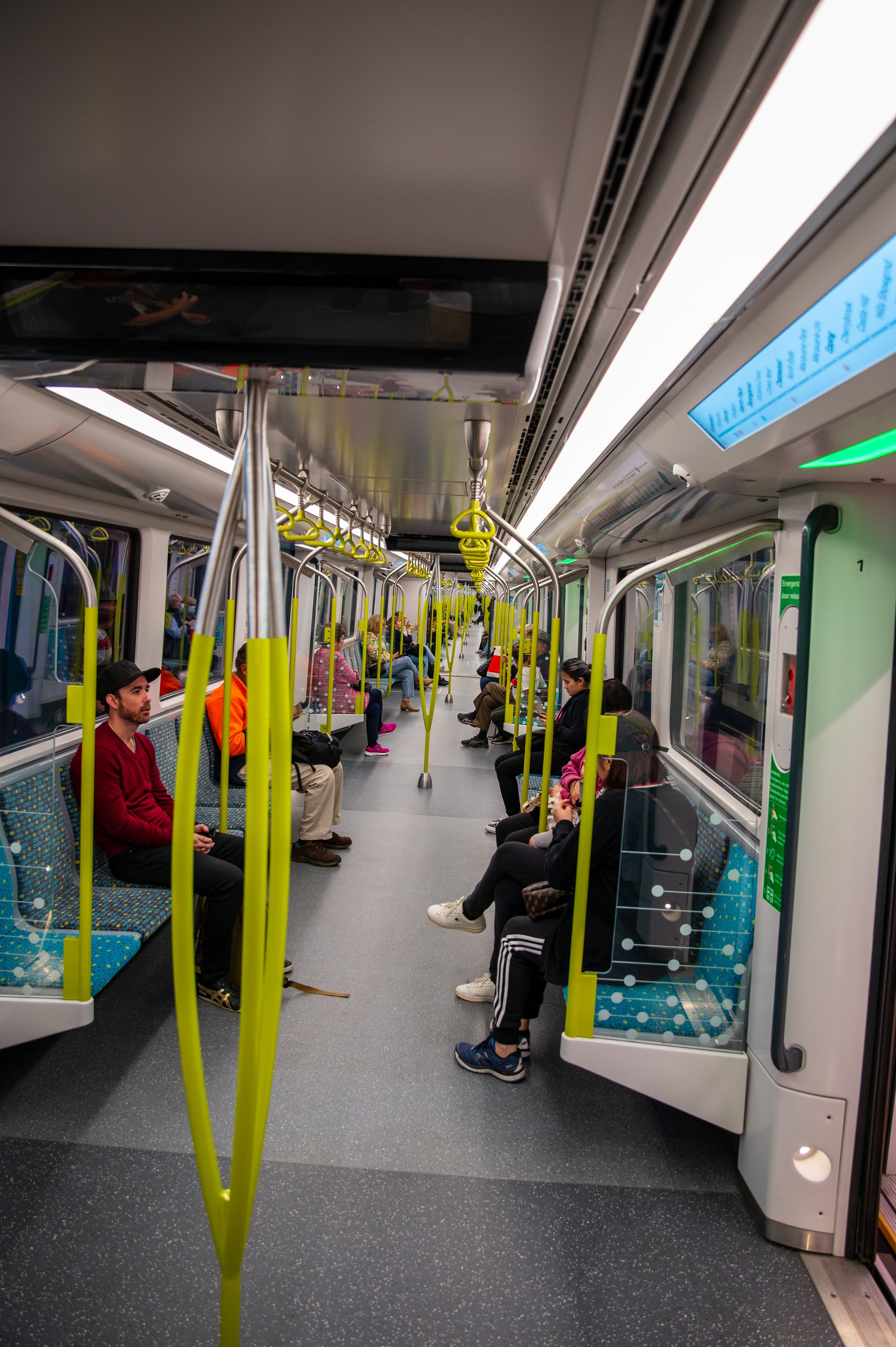 Inside a modern subway train with passengers seated and standing, yellow support handles hanging from the ceiling, large windows showing reflections, and an electronic display screen overhead.