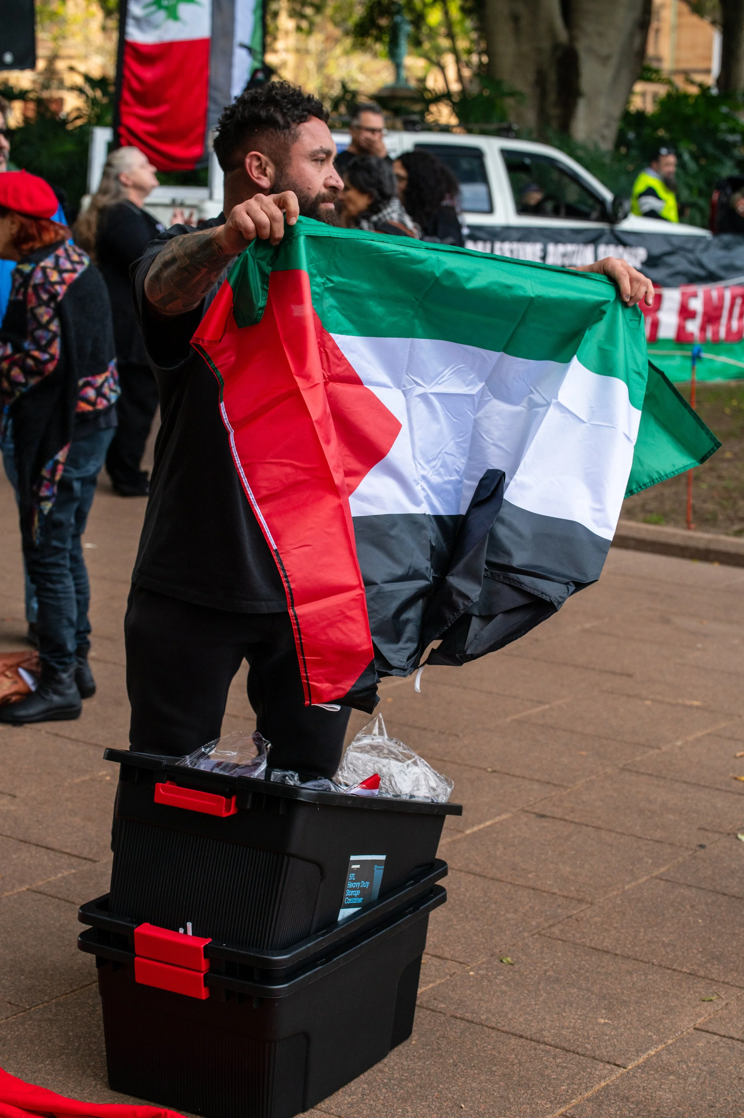 A man holding a Palestinian flag at a public demonstration or rally, with other people and banners in the background.