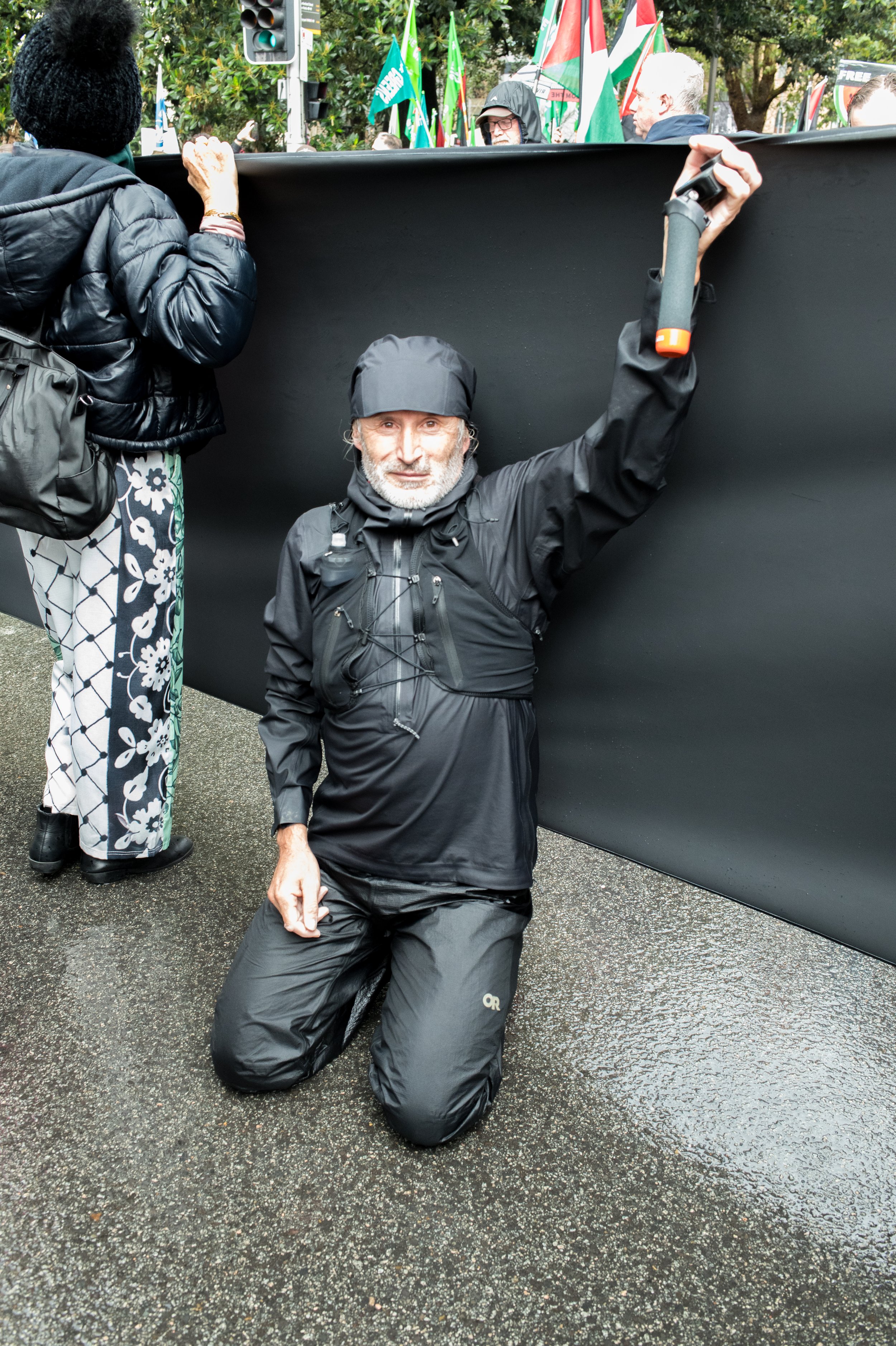 A man in black outdoor gear kneeling on wet pavement, holding a black barricade with his right hand during a protest or march.