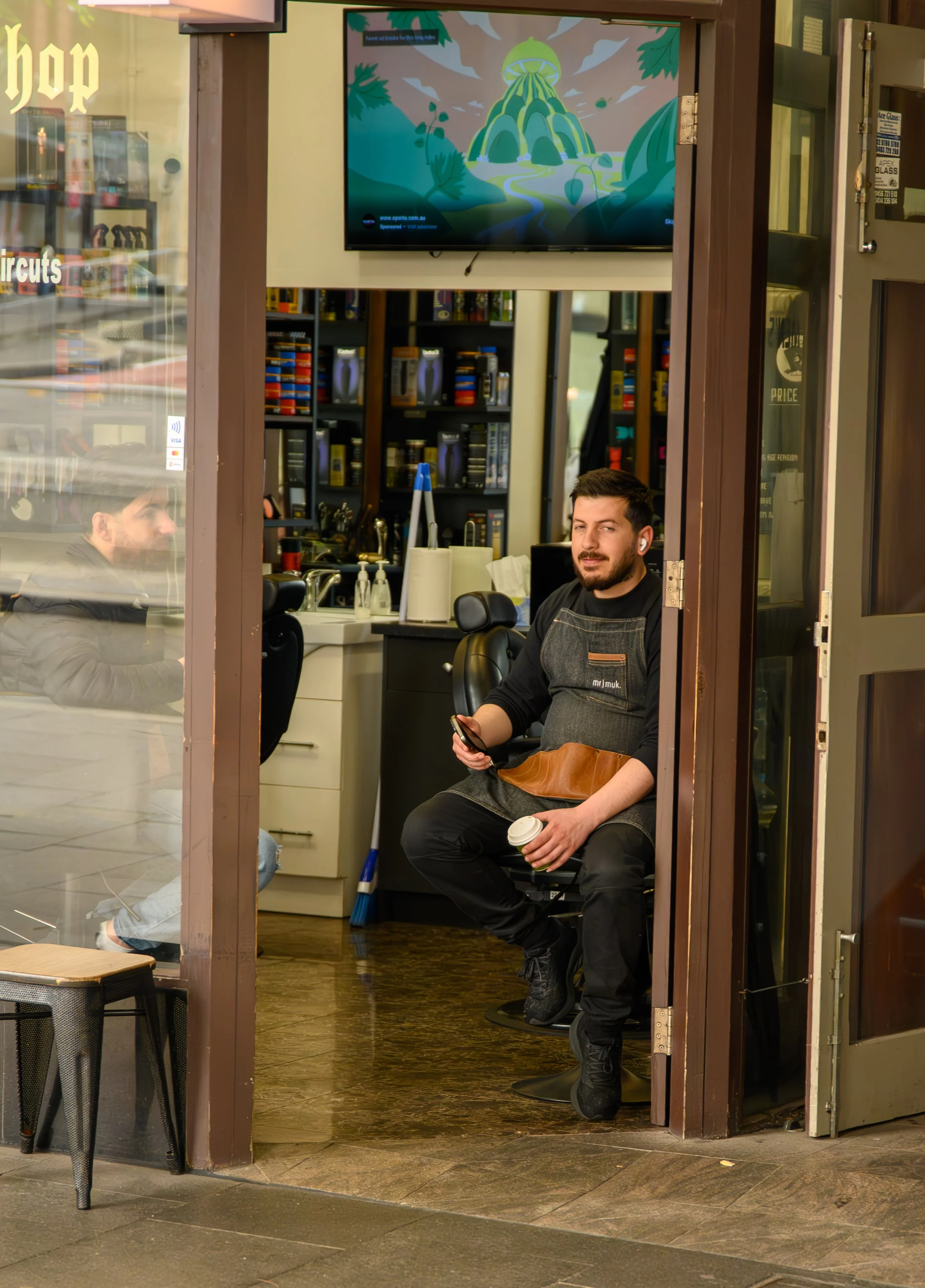 Barber sitting in a chair at his station in a barbershop, holding a coffee cup and a phone, with a mirror, shelves, and a television in the background.