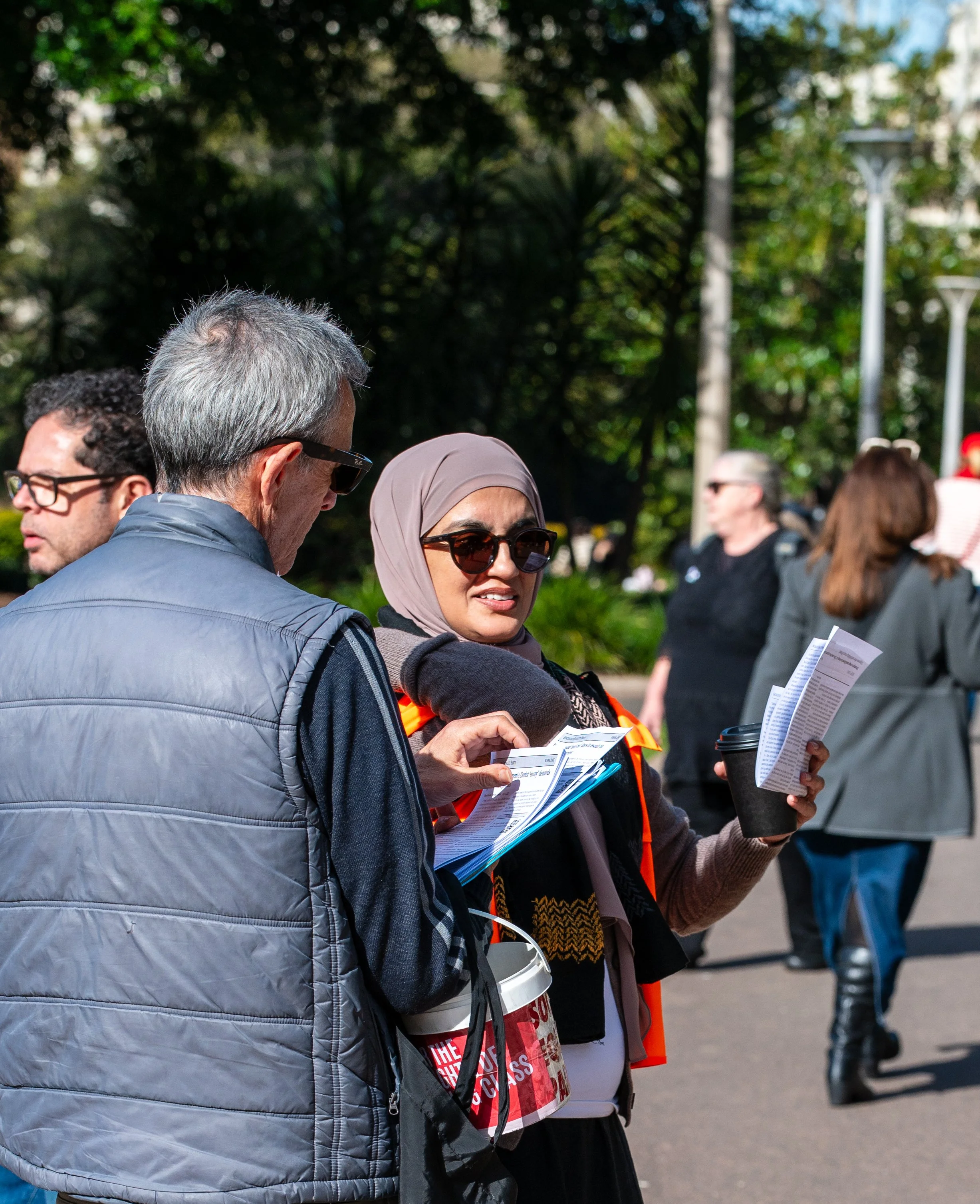 Two people, a man and a woman, standing outdoors, reading pamphlets or flyers. The woman is wearing a hijab and sunglasses, and the man has gray hair and sunglasses. There are other people in the background and trees, indicating a public park or similar setting.
