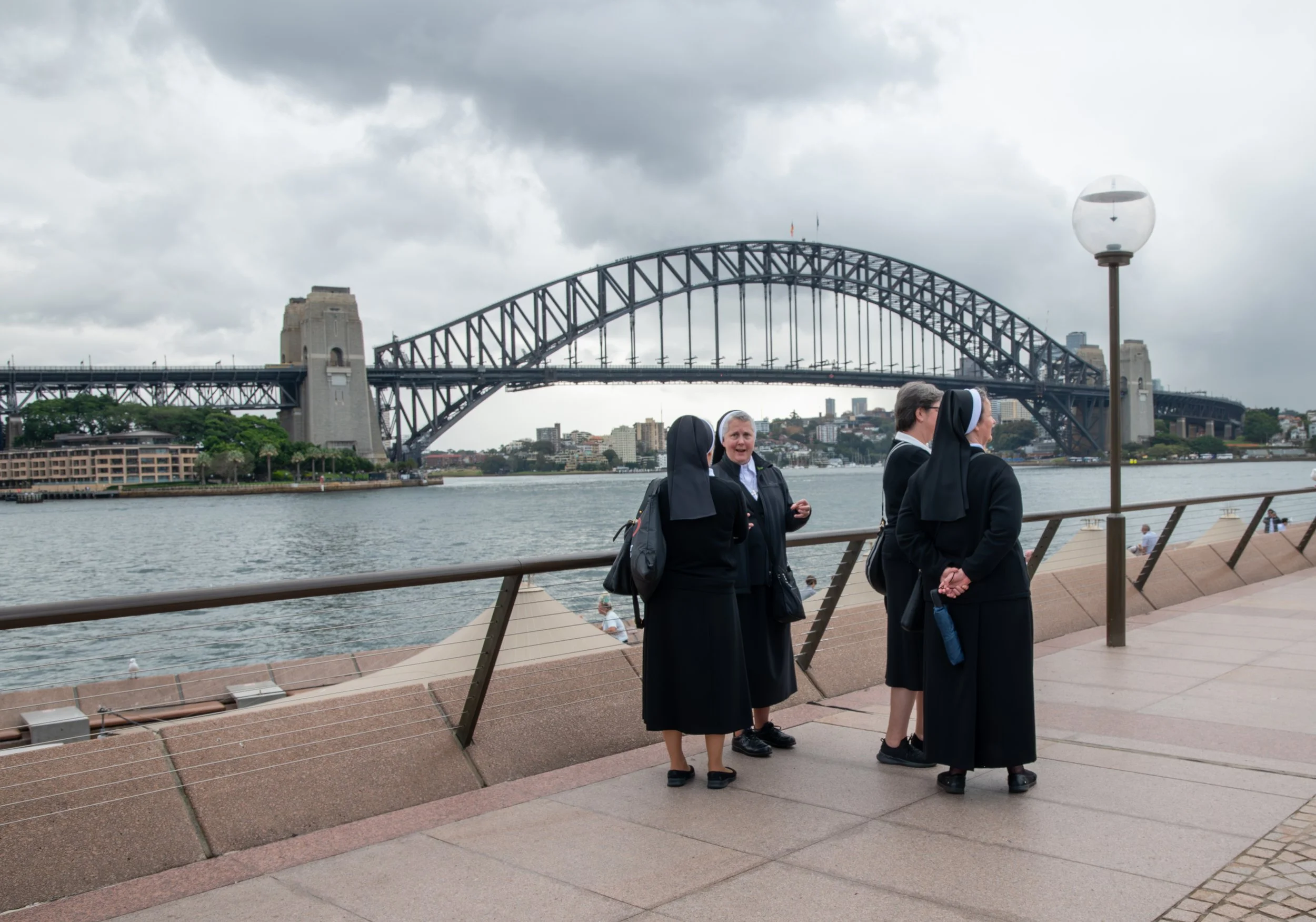 Four nuns in black habits standing by Sydney Harbour, with the Sydney Harbour Bridge in the background under a cloudy sky.