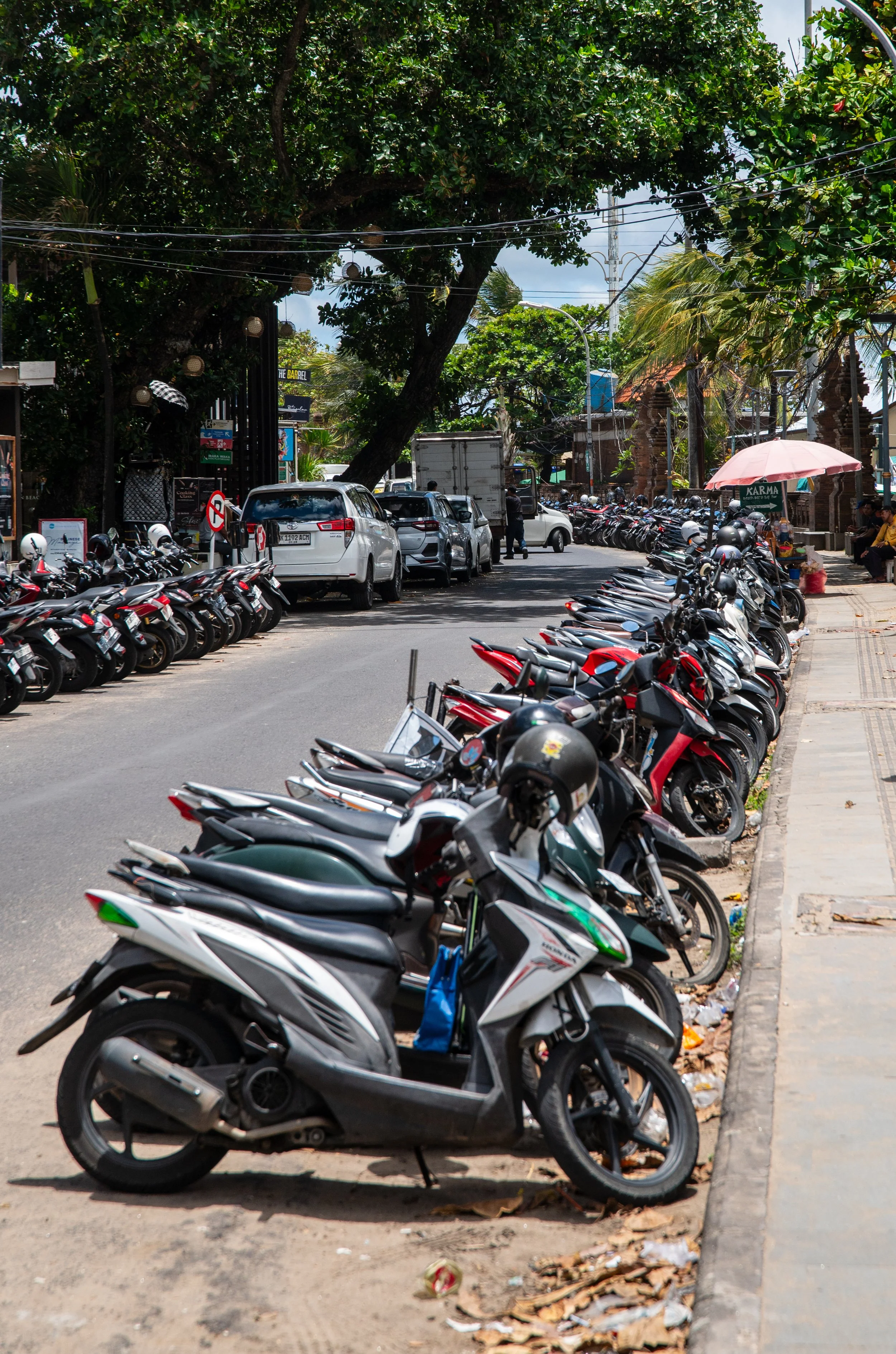 Parking area filled with numerous parked motorbikes along the curbside and a few cars driving down the street. Large trees provide shade over the scene, with a pink umbrella on the right side. There are buildings, signs, and electric wires in the background under partly cloudy skies.