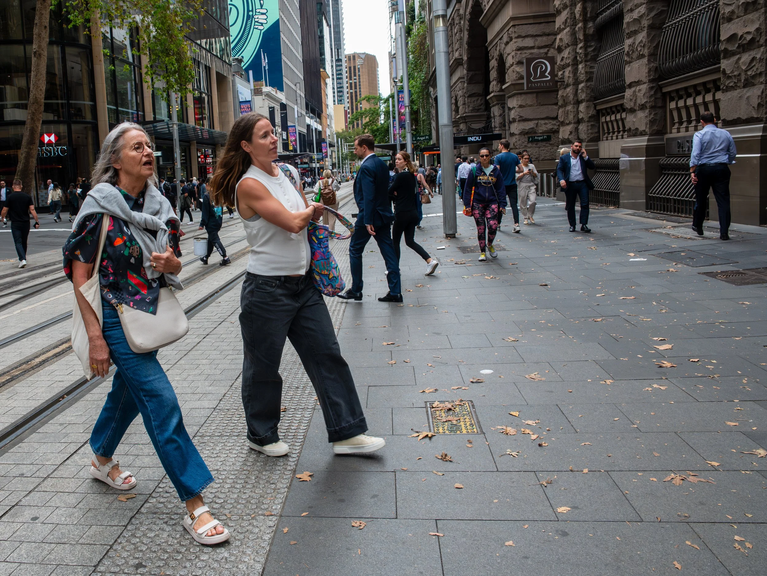 Crowded city street with pedestrians walking, including two women in the foreground, taller buildings, and streetcar tracks