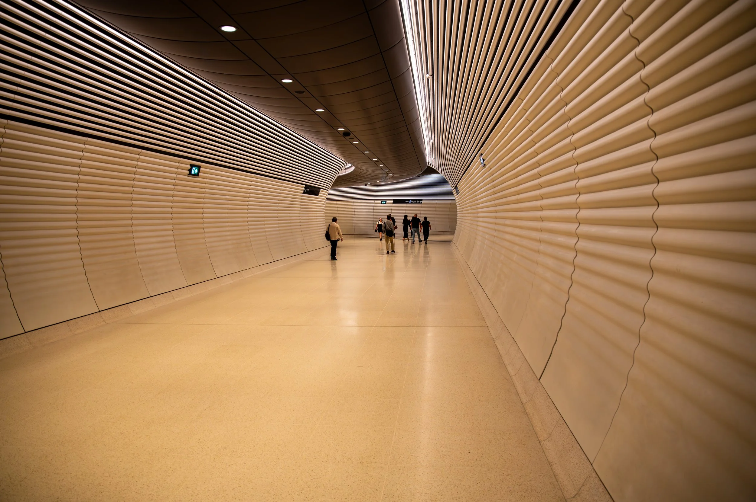 A modern, curved tunnel with beige and brown ribbed walls, ceiling, and floor, and a few people walking through it.