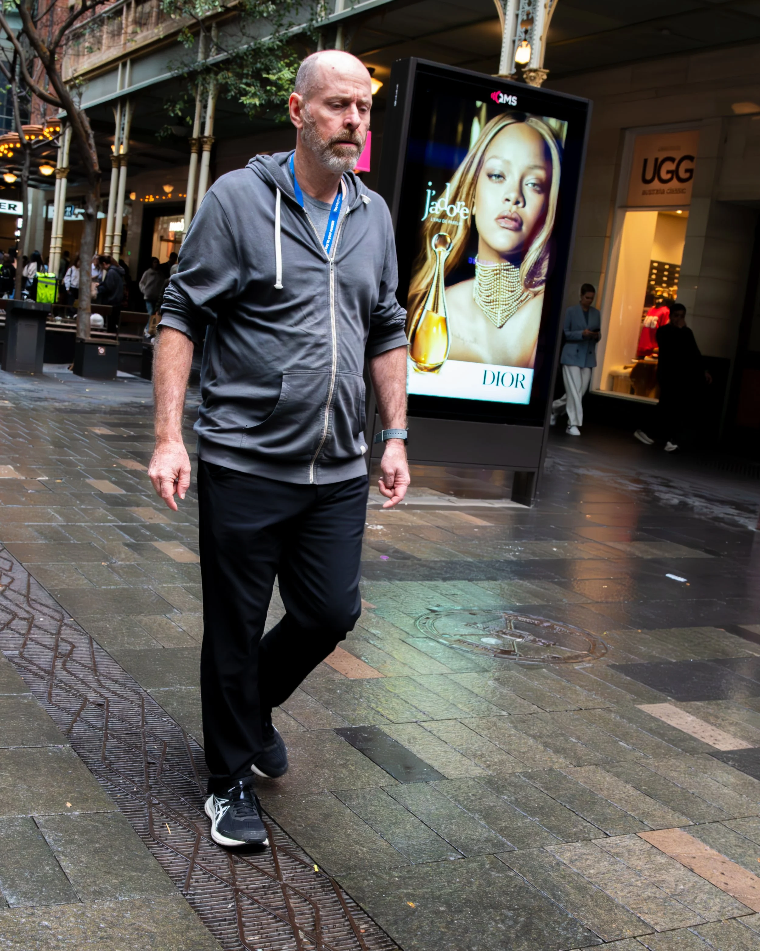 A man with a white beard wearing a gray hoodie and black pants walking on a wet city sidewalk near a digital billboard displaying an advertisement for Dior. The sidewalk is wet, indicating recent rain, and there are other pedestrians and storefronts in the background.