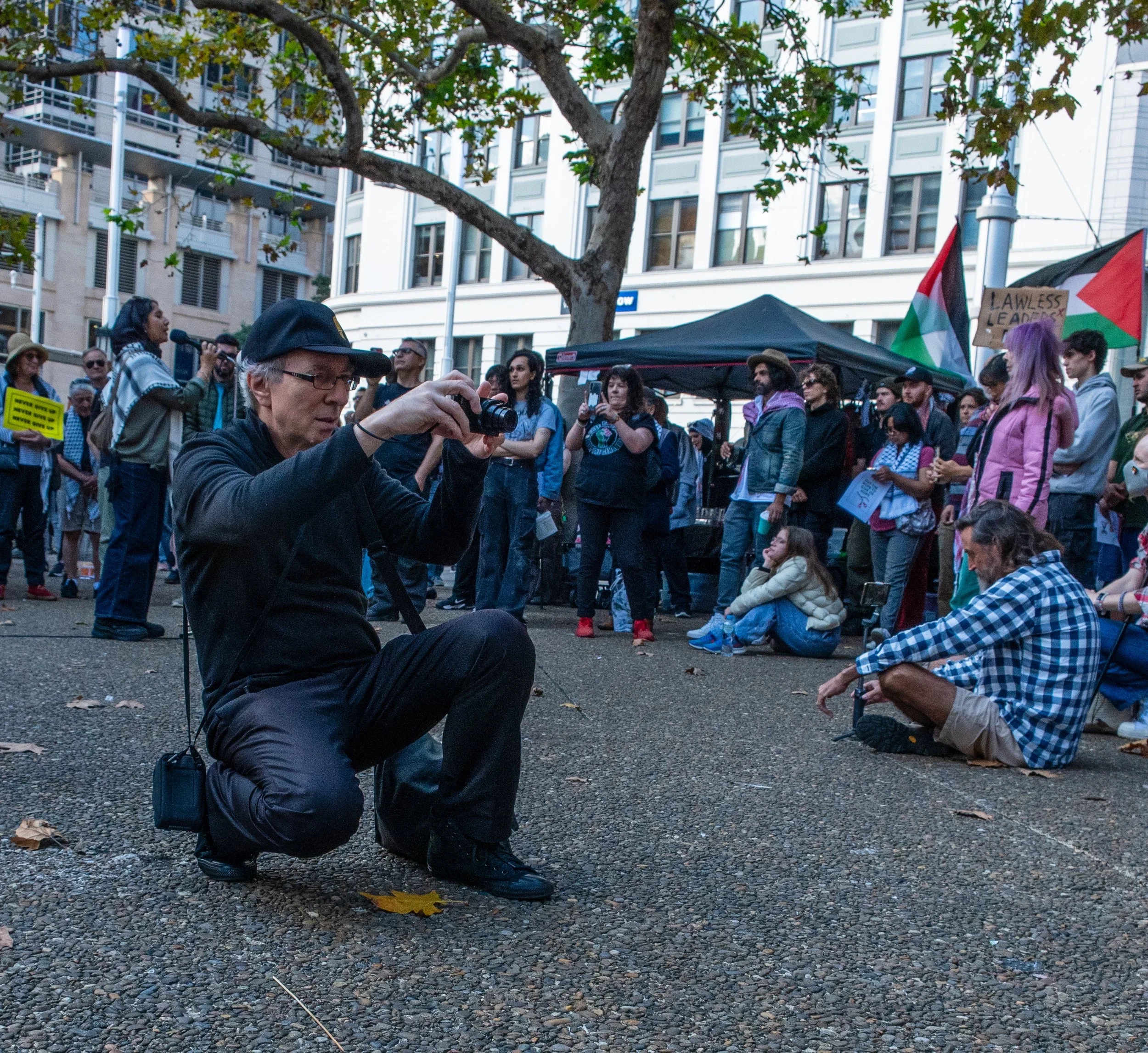 A man wearing a black cap and dark clothing kneels to take a photo at a protest or rally. Behind him, a crowd of diverse people are gathered, some holding signs and flags, in an urban setting with buildings and trees.