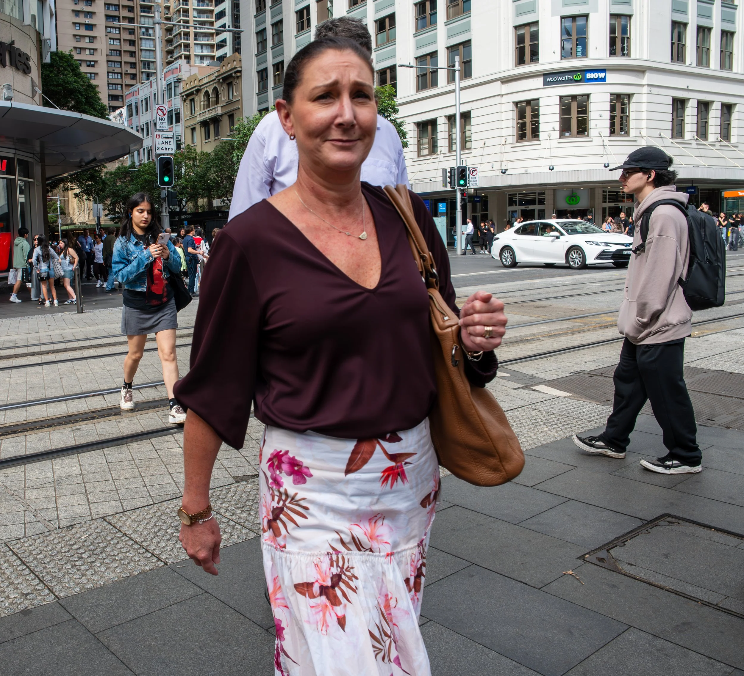 A woman with dark hair tied back, wearing a dark purple top and a white skirt with a pink and brown floral pattern, walking in an urban street scene with buildings, cars, and other pedestrians.