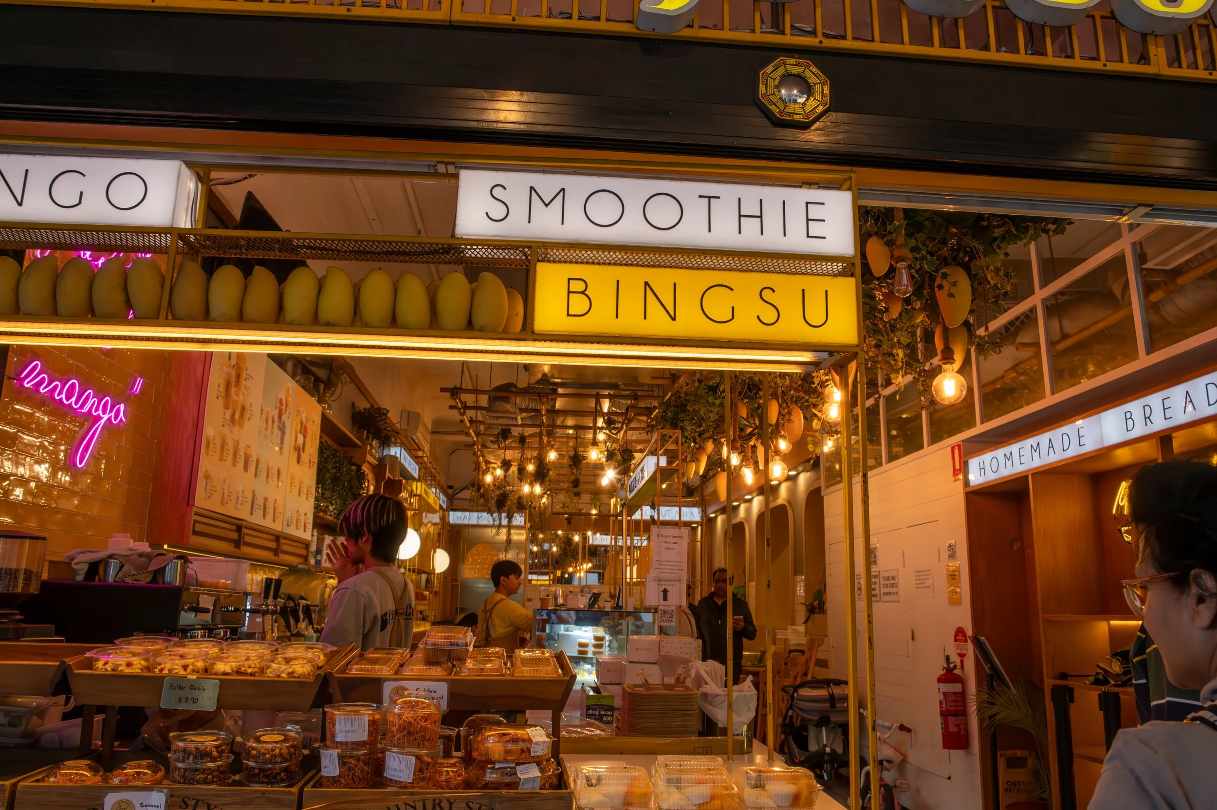 Interior of a food stall with illuminated signs for smoothies and bingsu, display of fresh fruits, baked goods, and customers inside.