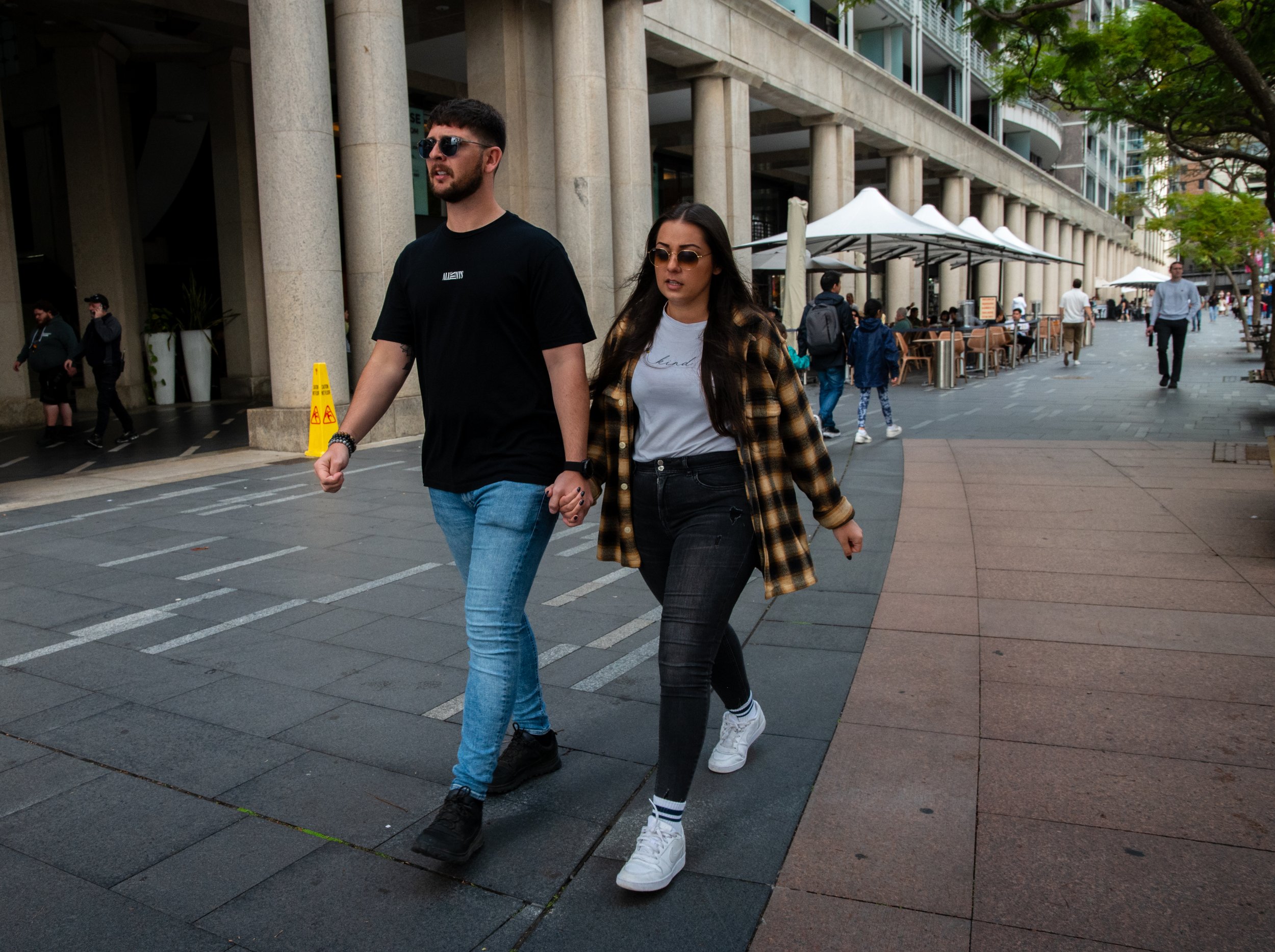 A man and woman walk hand-in-hand down a city sidewalk, with outdoor seating and buildings in the background.