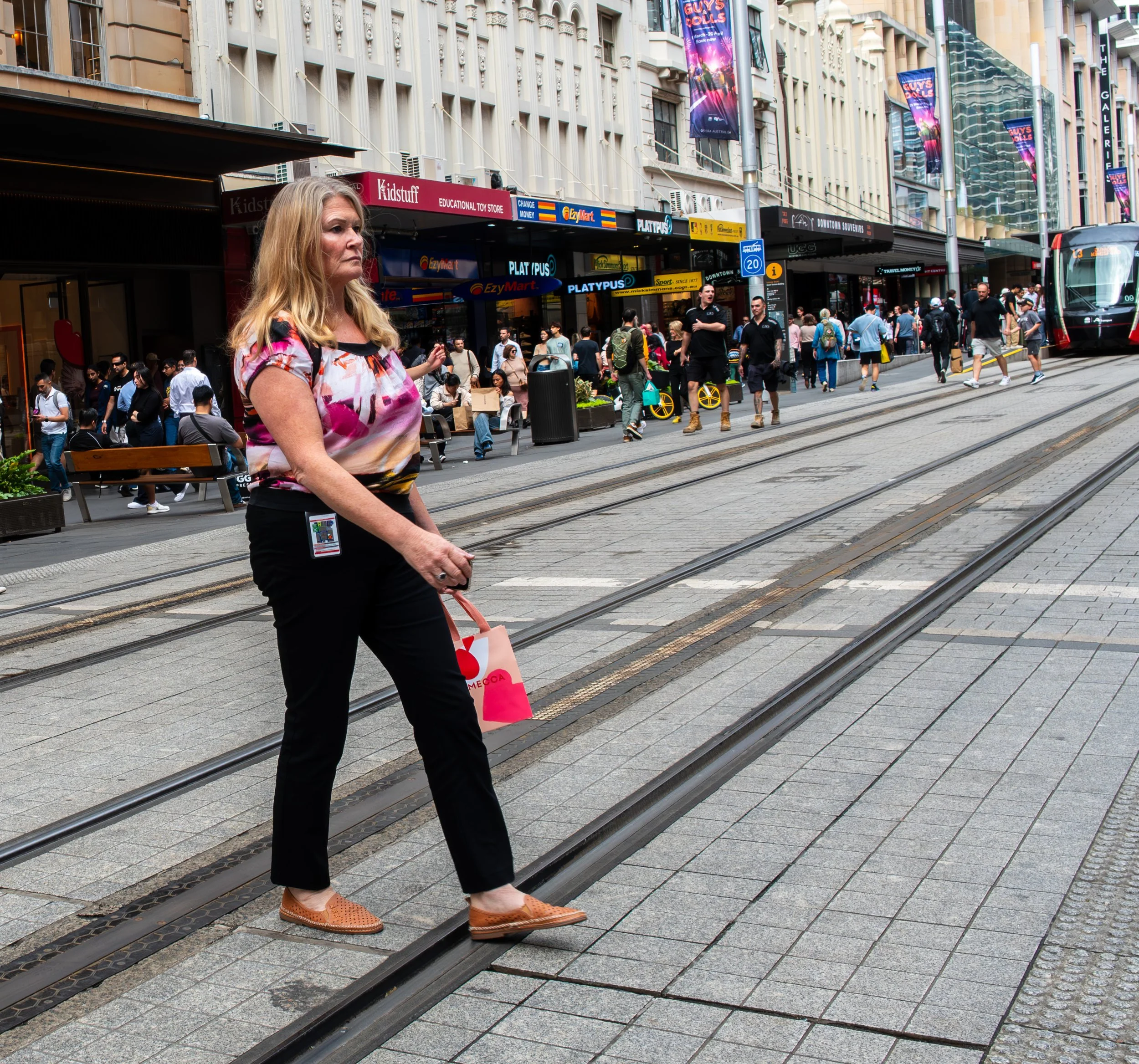 A woman crossing a busy city street near tram tracks, holding a pink shopping bag, with shops and a crowd of pedestrians in the background.