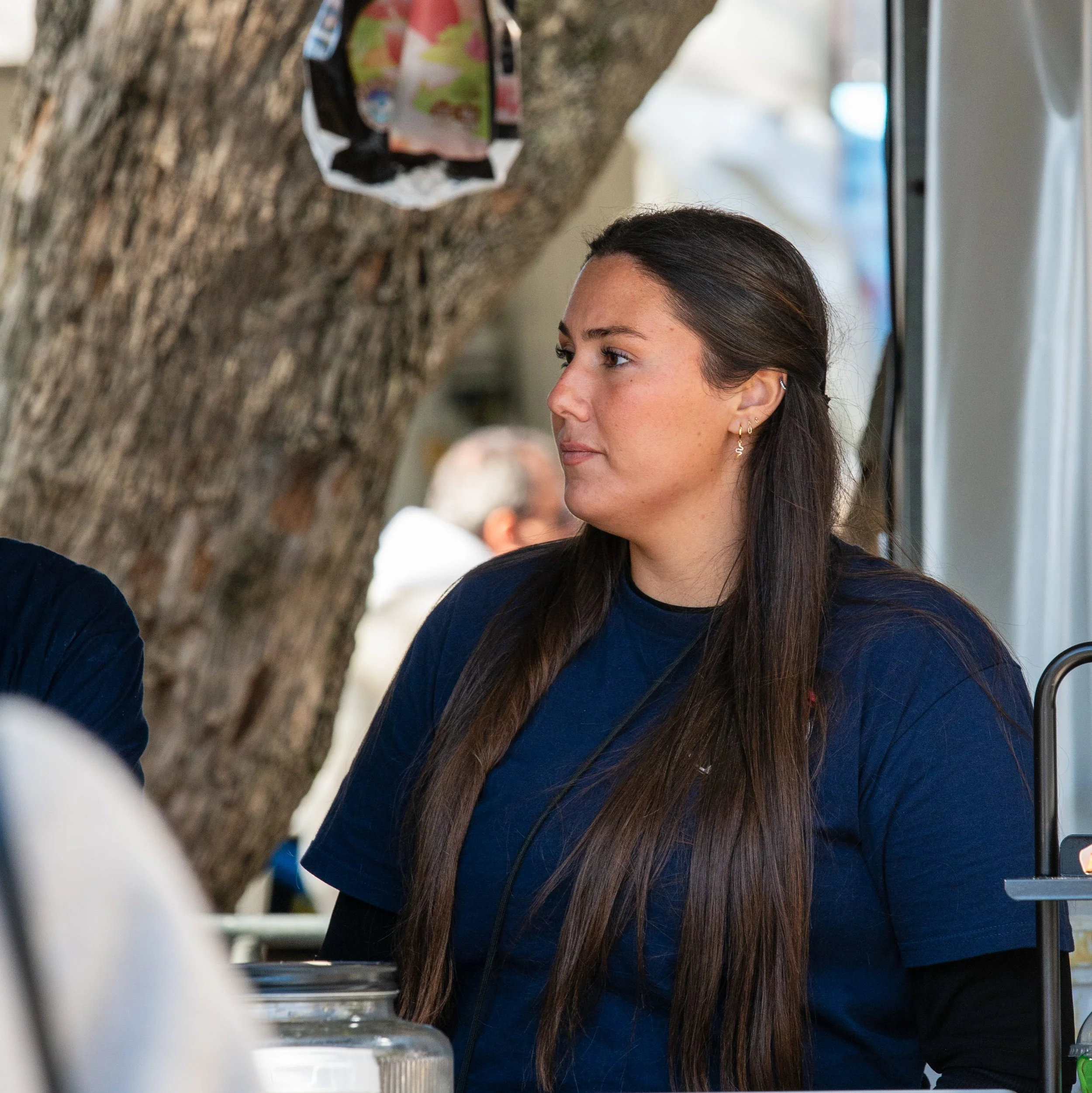 Young woman with long dark hair and small earrings, wearing a navy shirt, standing near a tree with a food bag hanging on it, indoors.