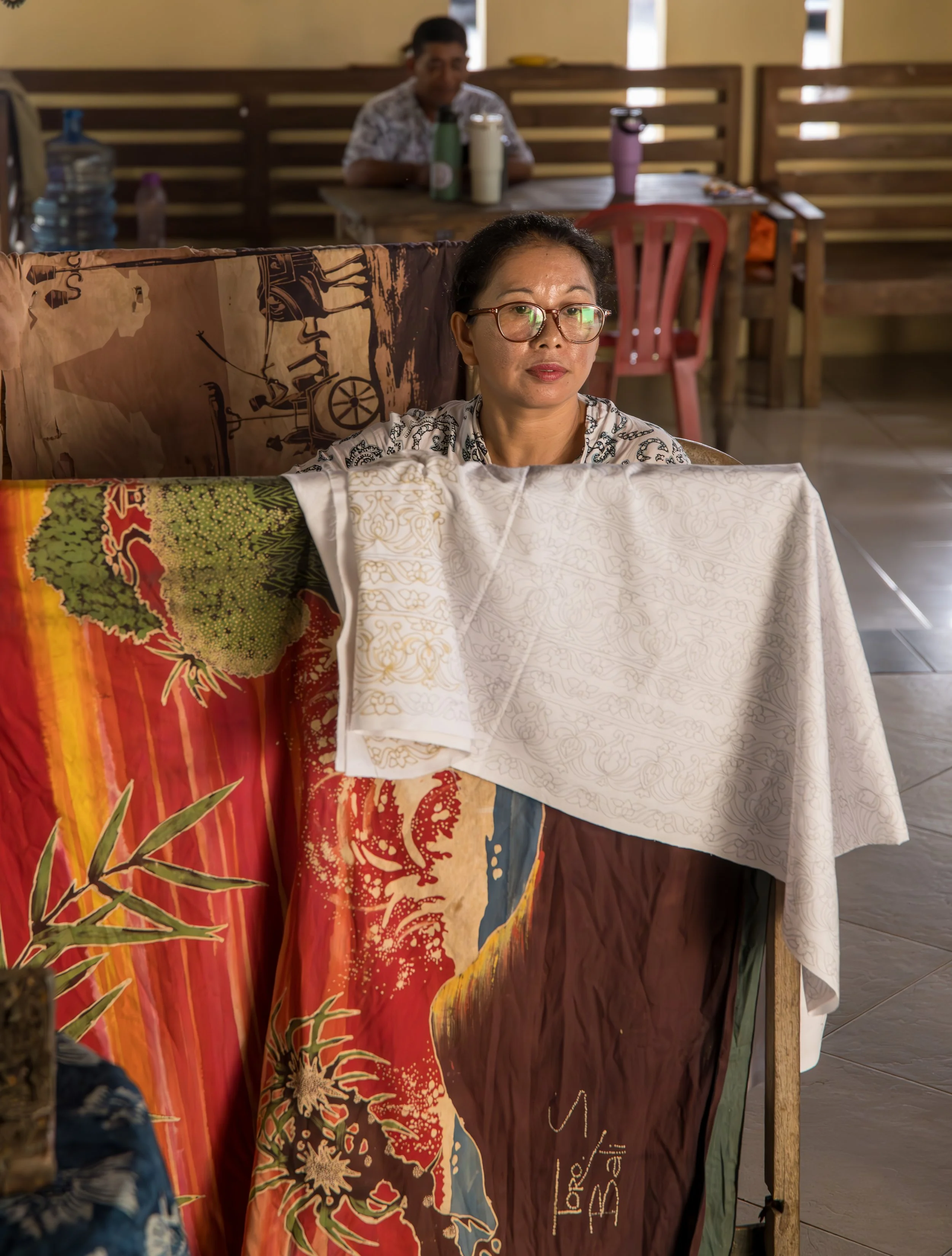 A woman with glasses sitting behind a cloth with vibrant patterns, inside a room with wooden furniture and another woman in the background at a table.