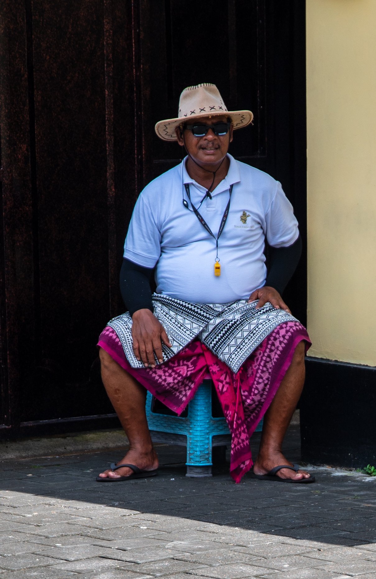 A man sitting on a blue plastic stool in front of a dark wooden wall and a yellow wall, wearing a straw hat, sunglasses, a white shirt, a patterned sarong, and flip-flops, with a whistle hanging around his neck.