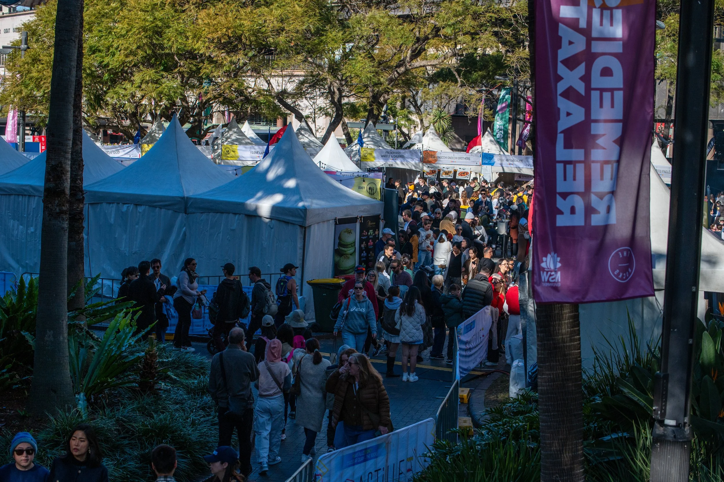 Crowd of people walking through an outdoor festival with white tents and colorful banners.