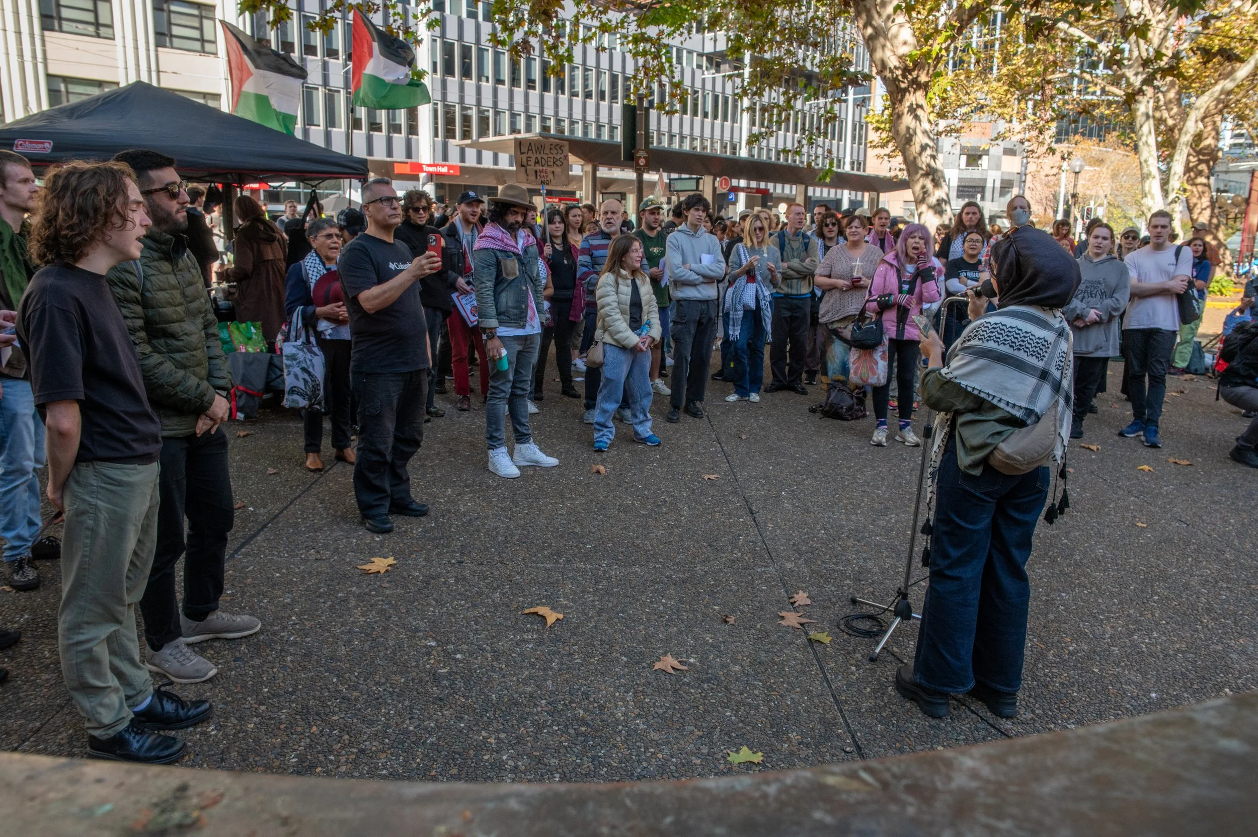 A woman with a headscarf speaks into a microphone at a protest rally in a city park, with a crowd of people listening and holding signs and flags. The crowd is diverse, with some of them taking photos and others watching attentively.