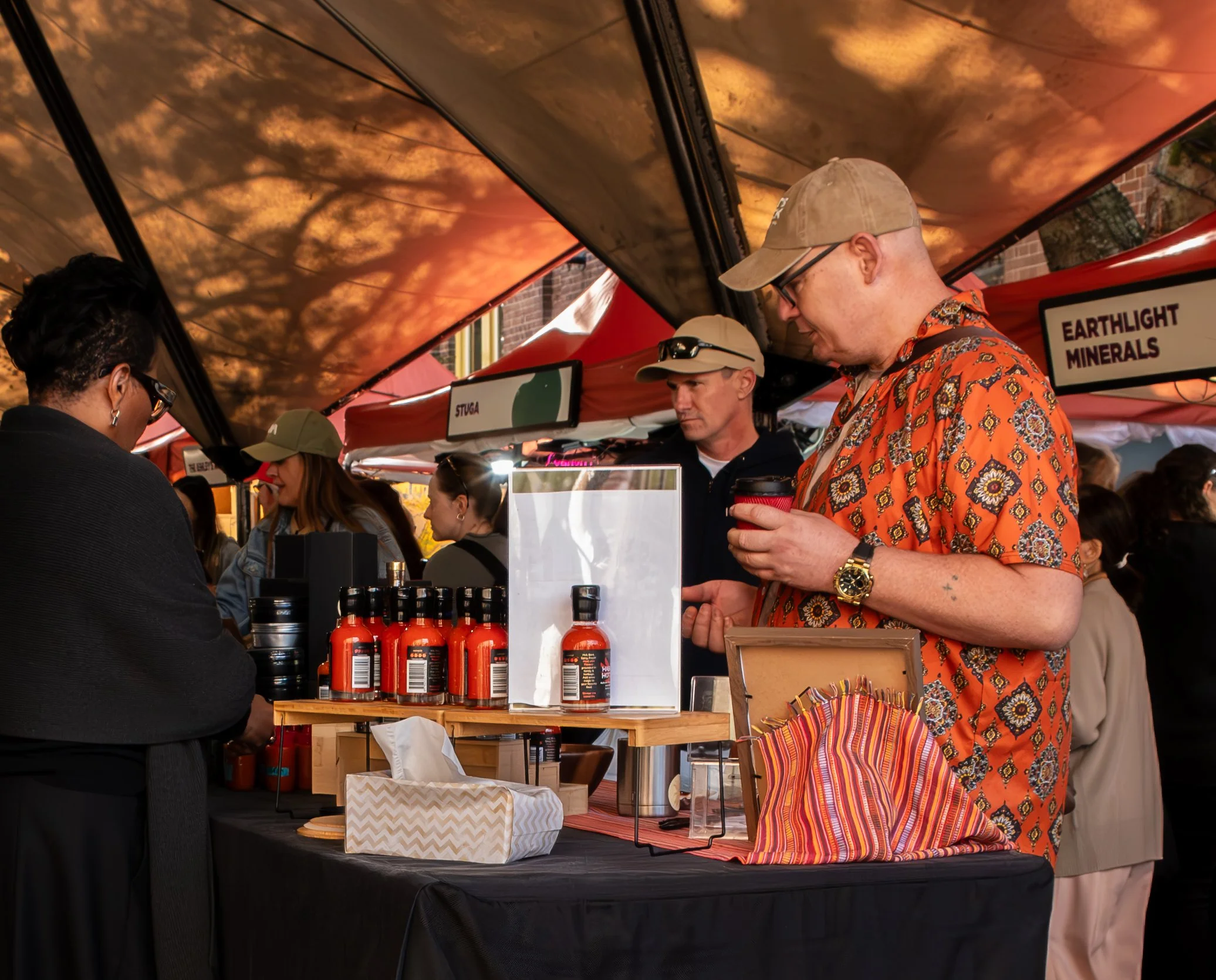People shopping at a market stall with condiments and minerals, under an orange, cloudy sky-themed canopy.