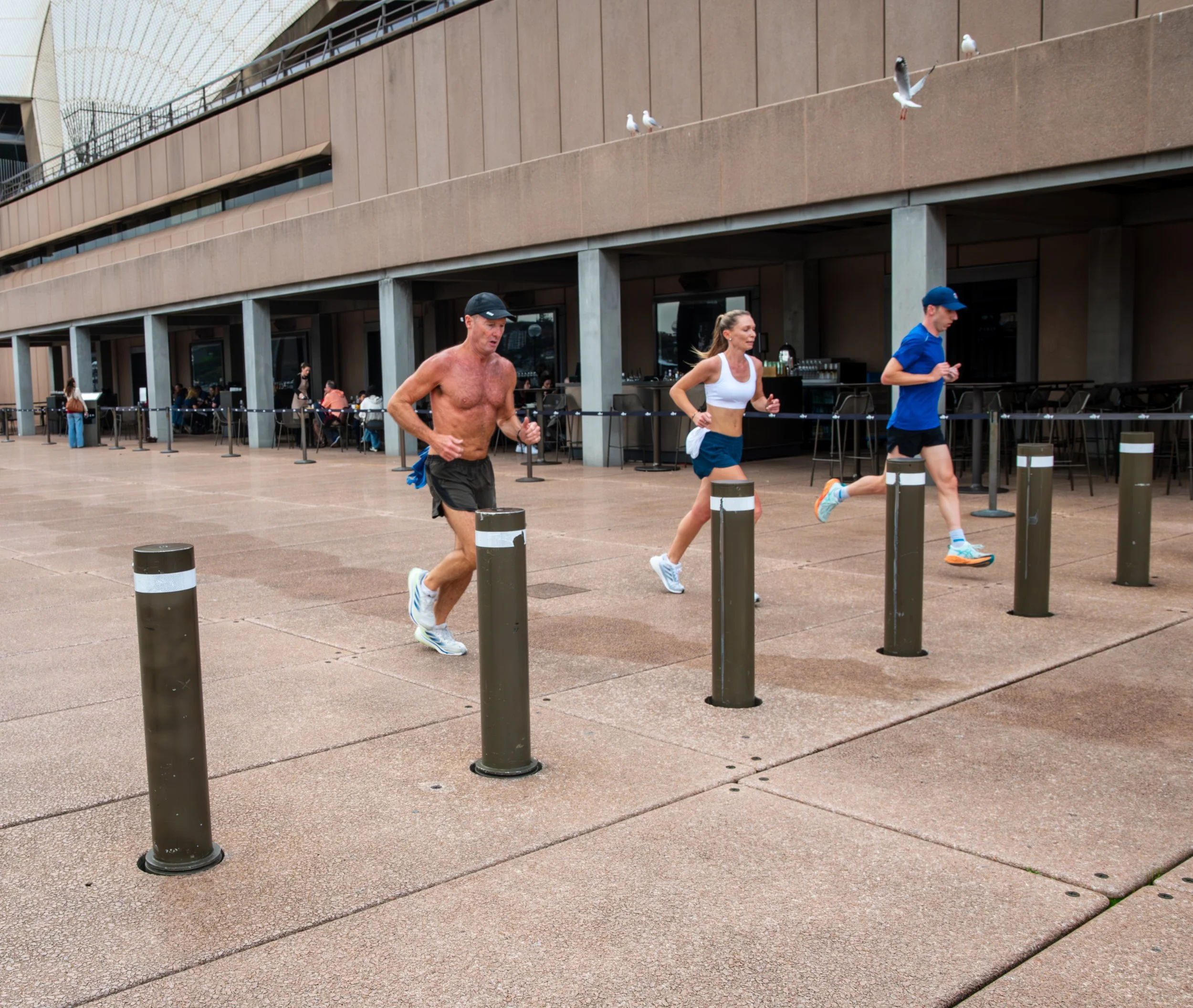 Three people running outdoors near a building with seated diners in the background, two men and a woman, with the shirtless man in front.
