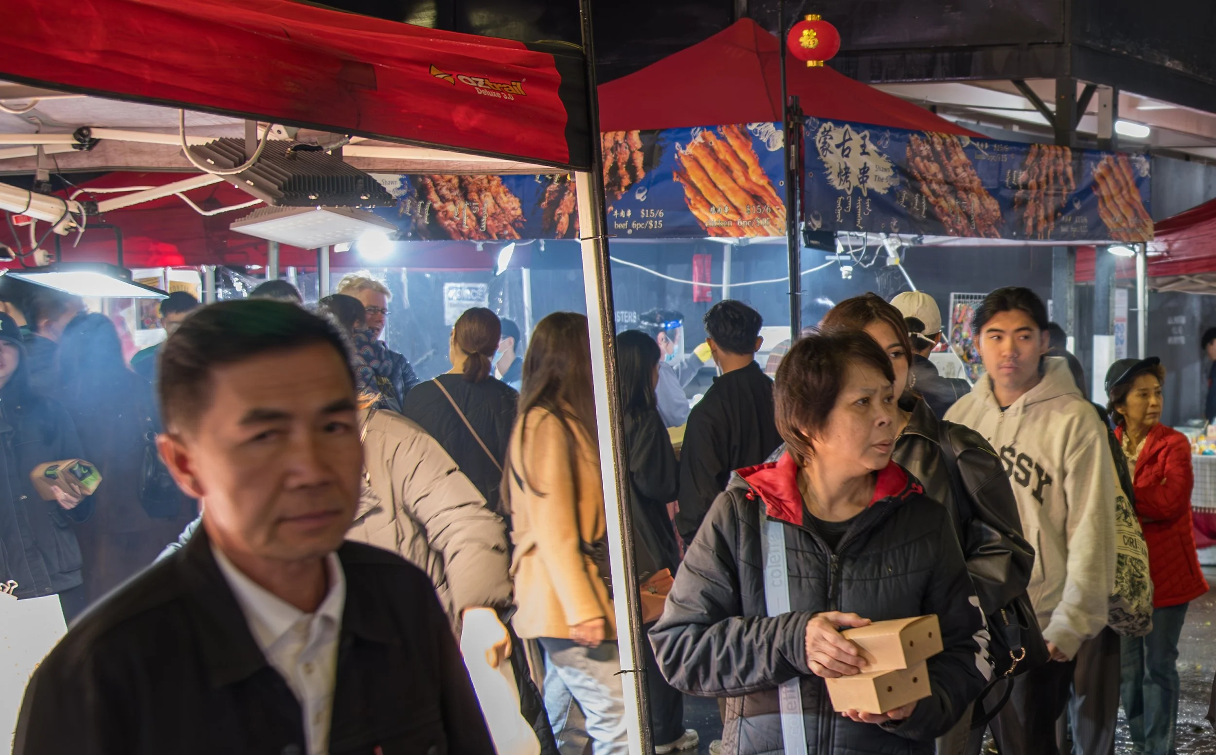 Crowded night market with people shopping at food stalls with red and blue signs, some people holding food items, illuminated by bright lights.