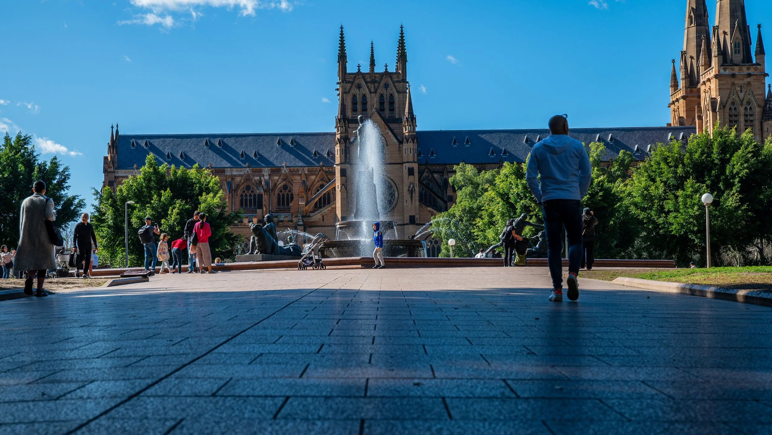 People walking and standing near a fountain in front of a large, Gothic-style cathedral with green trees and a blue sky in the background.