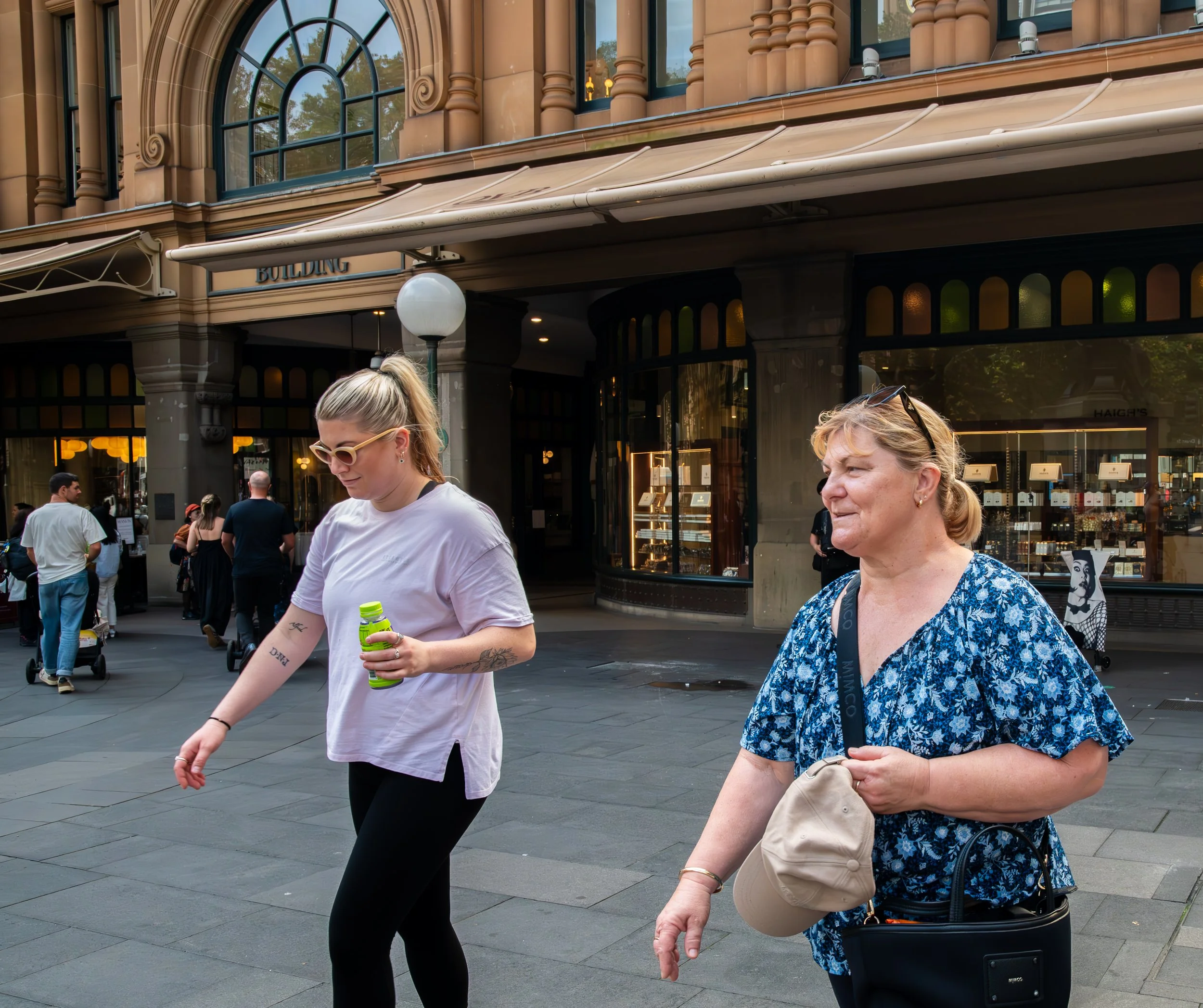 Two women walking on a city sidewalk in front of a building with large windows and decorative architecture.